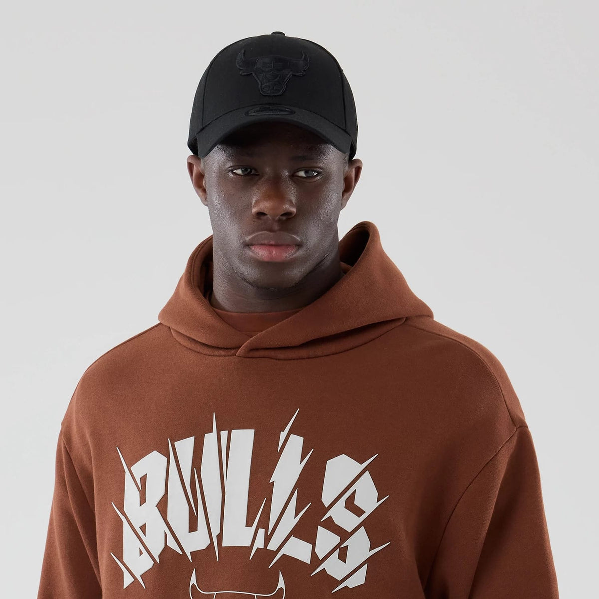 New Era Chicago Bulls NBA Core Plus Brown Pullover Hoodie, front view on-model showing the brown fabric and white Bulls lettering across the chest, paired with a black cap featuring an embossed Bulls logo at the centre front.