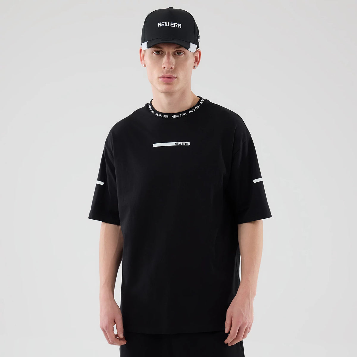 New Era Tech Black T-Shirt, front view on-model showing a fitted design with a white printed New Era logo across the chest, matching text along the collar edge, and thin white accents on the sleeves, paired with a black cap.