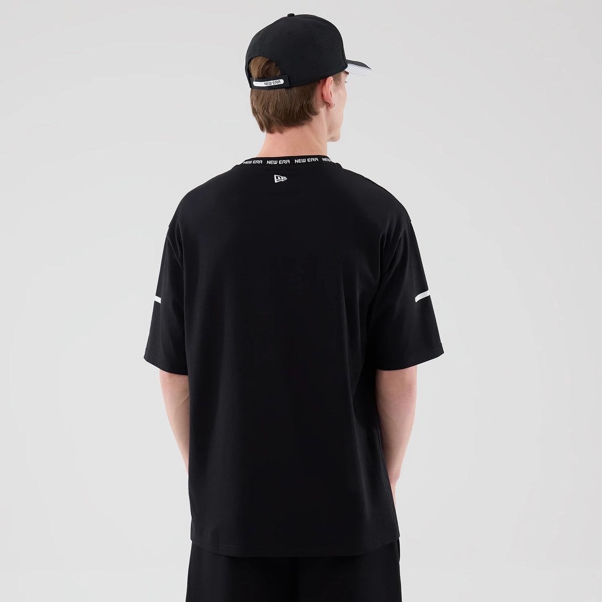 New Era Tech Black T-Shirt, back view on a model wearing a matching black cap, showing the New Era logo embroidered at the centre of the back near the neckline and contrasting white stripes on each sleeve near the elbow.