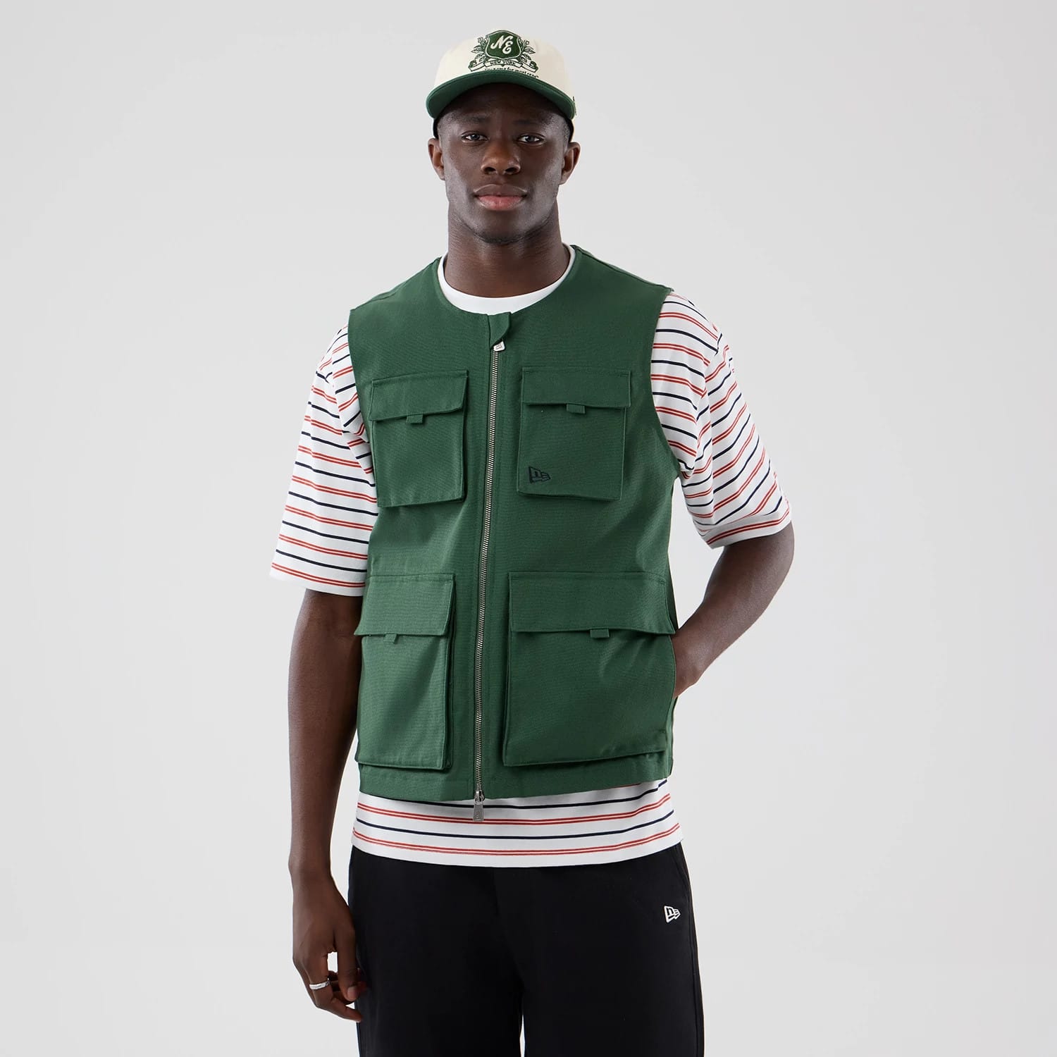 New Era Dark Green Utility Vest, front view on a model with a striped short-sleeve shirt underneath, featuring multiple front pockets with flap closures and a central zip fastening.