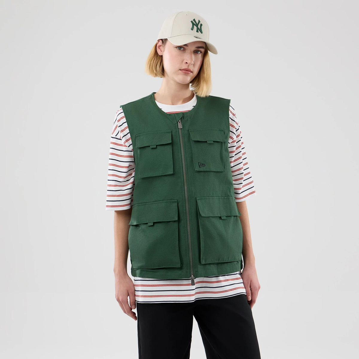 New Era Dark Green Utility Vest, front view on a model styled with a striped T-shirt and baseball cap, featuring a full-length front zipper, multiple flap pockets on the chest and waist, and an embroidered New Era flag logo on the left chest.
