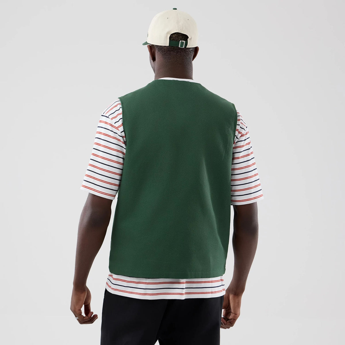 New Era Dark Green Utility Vest, back view on a model wearing a striped T-shirt and a cream cap with a dark green fastening, highlighting the vest’s dark green fabric and sleeveless design.