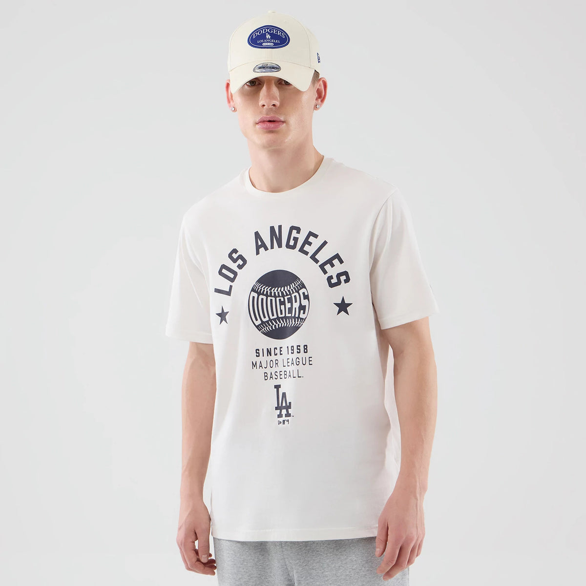 New Era LA Dodgers MLB Graphic Open White T-Shirt, front view on-model showing black Los Angeles lettering above a large baseball graphic, with additional text and a classic LA logo below, paired with a white cap featuring a blue embroidered patch.