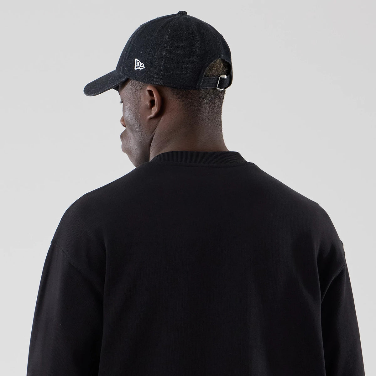 New Era Black Oversized Crew Neck Sweater, rear view on a model with a dark-coloured cap, showing the relaxed fit and ribbed neckline of the sweater, complementing the cap with a New Era flag logo on the left side.