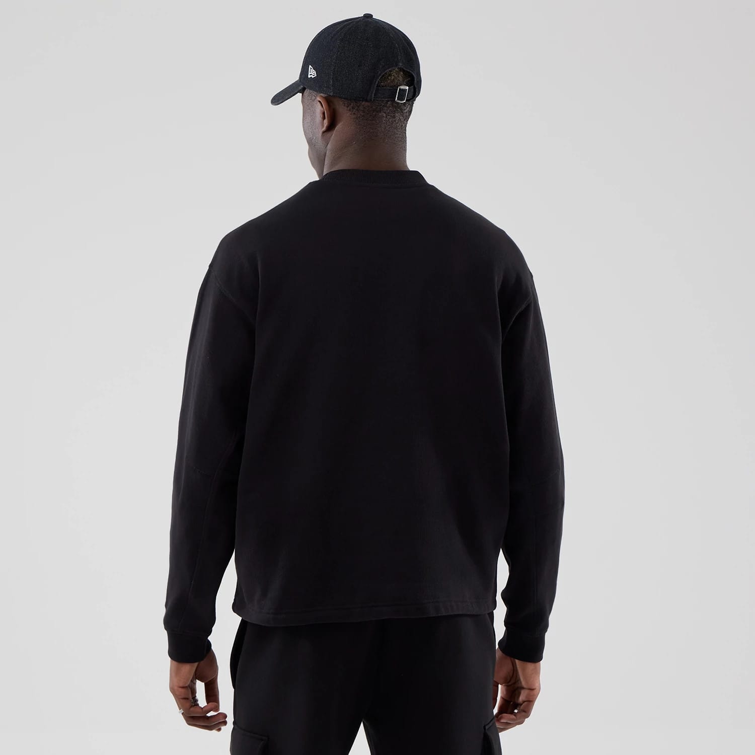 New Era Black Oversized Crew Neck Sweater, back view on a model wearing a black cap, showing the simple design with long sleeves and ribbed cuffs, paired with casual black trousers.