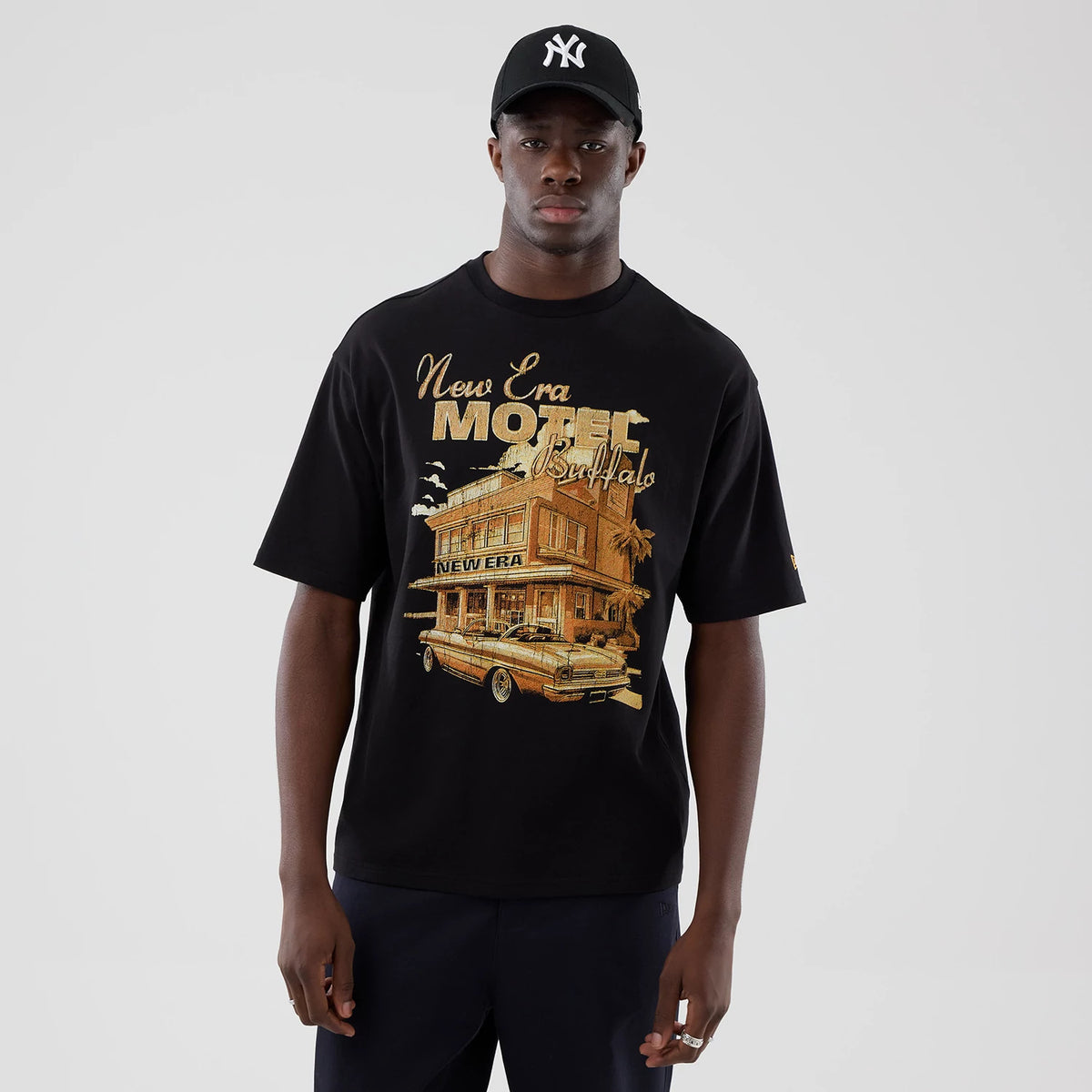 New Era Motor Graphic Black Oversized T-Shirt, front view on-model featuring a large golden motel print with 'New Era Buffalo' text across the chest, short sleeves, and a small embroidered New Era flag logo on the left sleeve.
