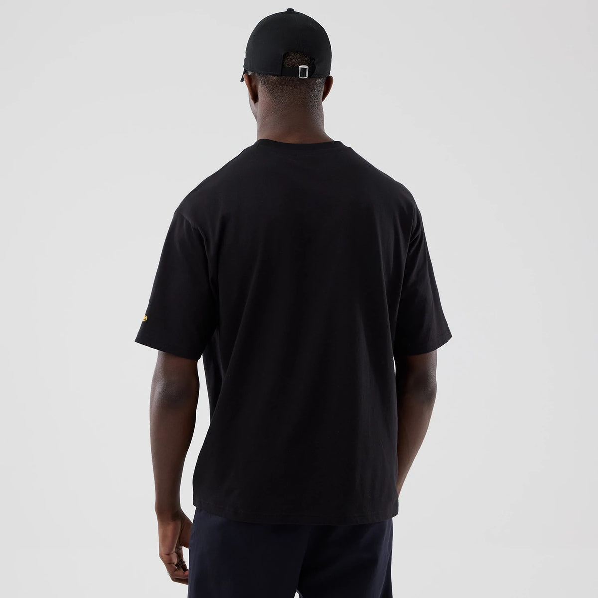 New Era Motor Graphic Black Oversized T-Shirt, back view on-model featuring a plain black back, short sleeves with a logo on the right sleeve, and paired with a black cap showcasing a fastening at the rear.