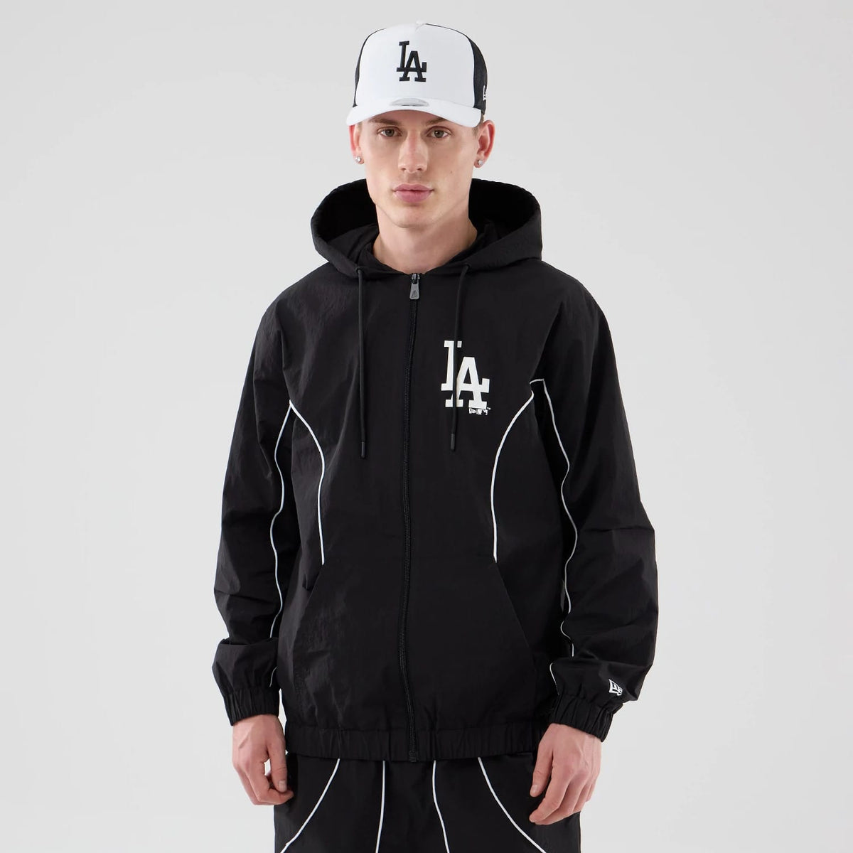 New Era LA Dodgers MLB Piping Black Jacket, front view on a model paired with a matching cap, showing a white embroidered LA logo on the left chest, white piping outlining the front, and an embroidered New Era logo on the left cuff.