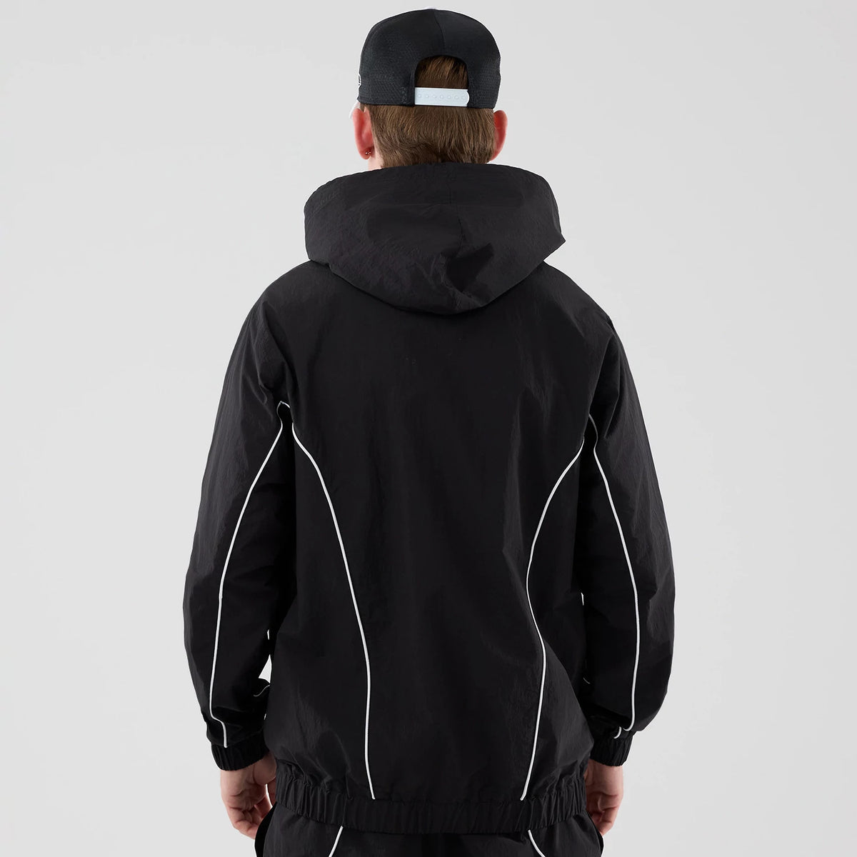 New Era LA Dodgers MLB Piping Black Jacket, back view on-model featuring a loose hood, white piping running along the shoulders and arms, and a cinched waistband, styled with a complementary dark cap with a light adjustable strap.