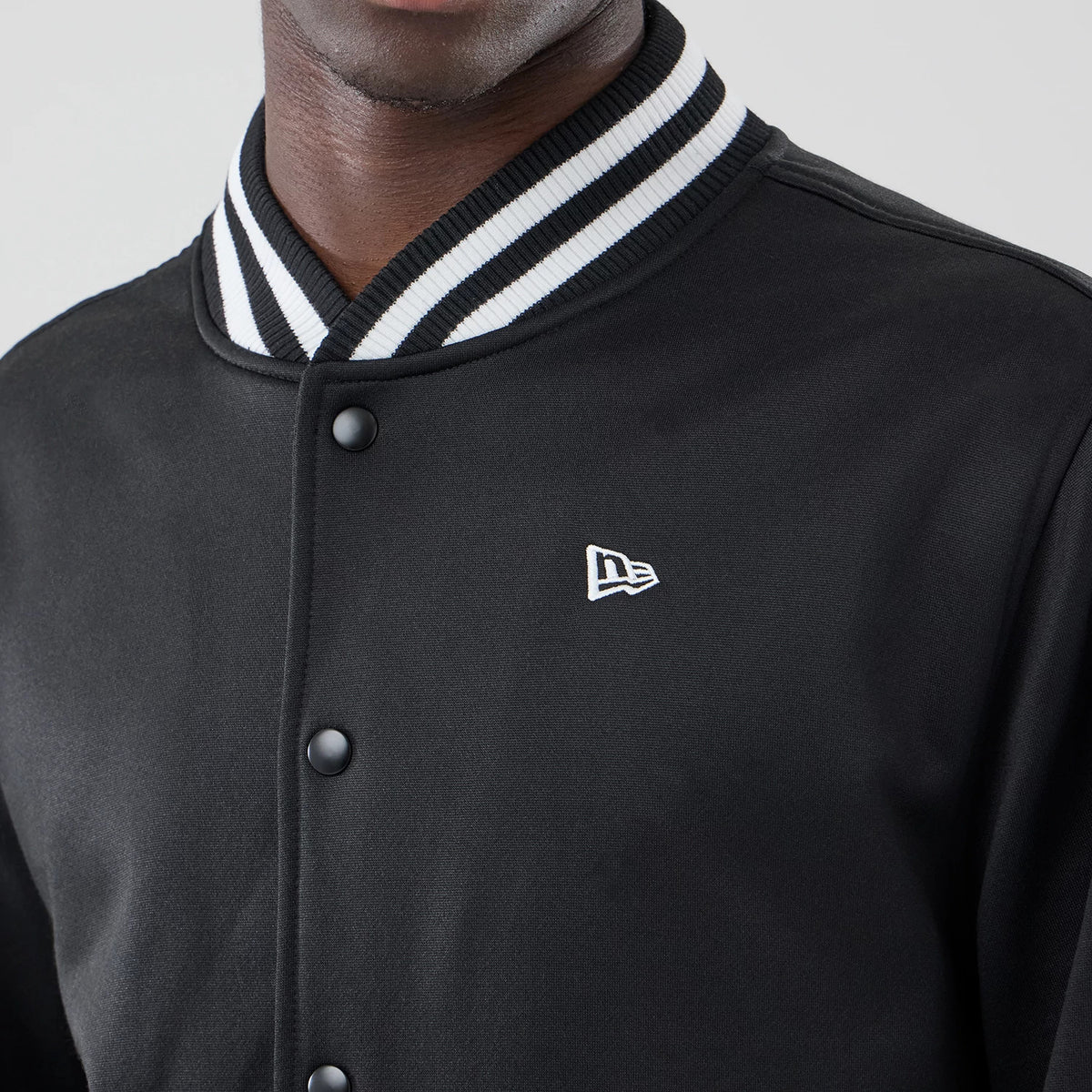 New Era Black Jersey Bomber Jacket, front view showing a white embroidered New Era logo on the left chest, black snap buttons down the centre, and a ribbed black-and-white striped collar, styled on a model.