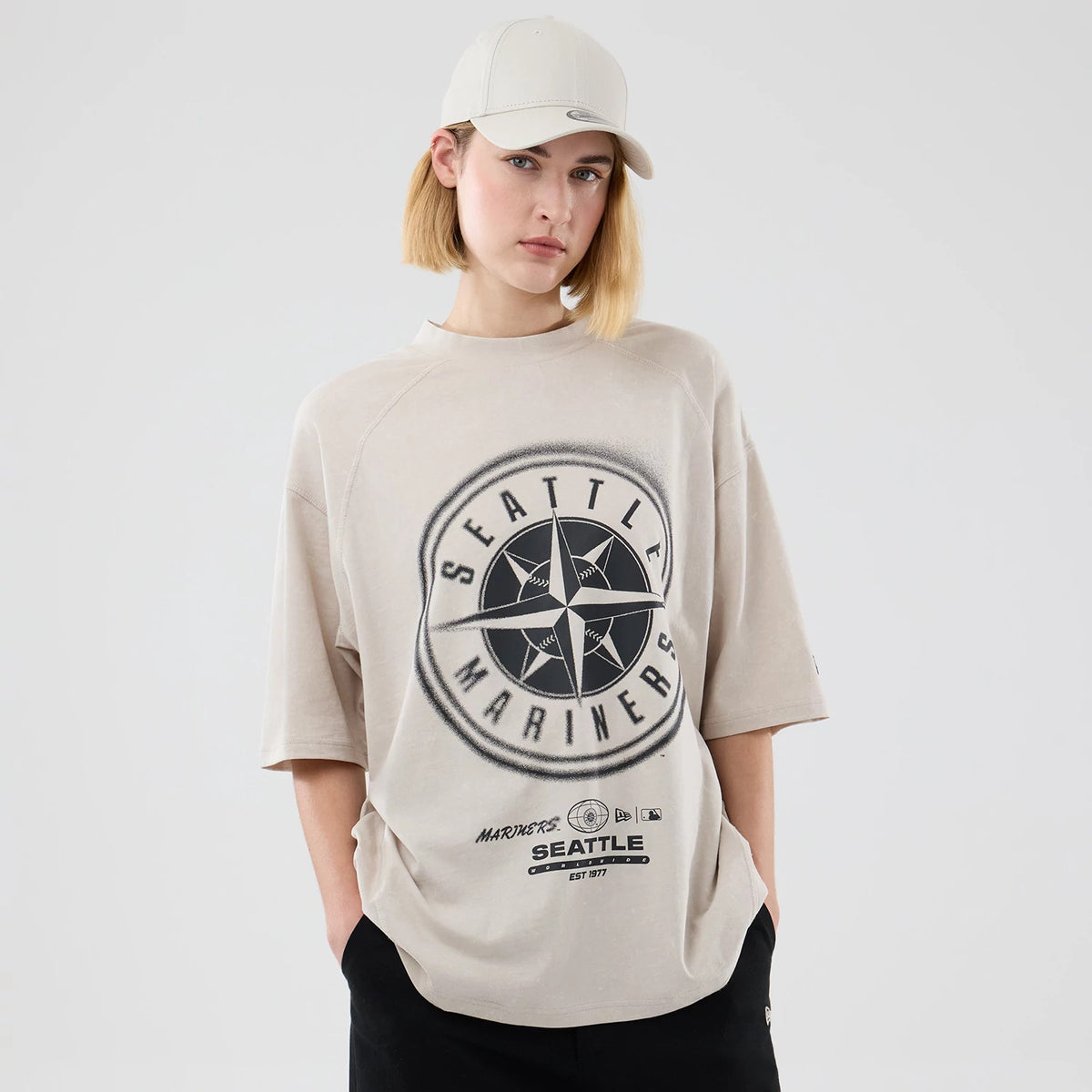 New Era Seattle Mariners MLB Graphic Cream Boxy T-Shirt, front view on a model with matching light cap, showcasing a large black graphic emblem on the chest featuring Seattle Mariners lettering and star design, with additional text and logos beneath.