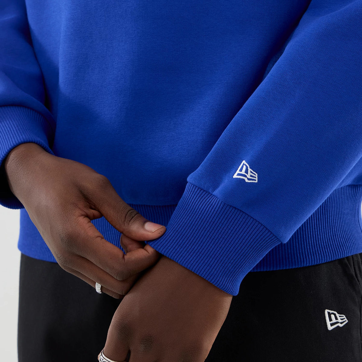 New Era Red Bull Racing Blue Oversized Crew Neck Sweater, close-up view showing the New Era flag logo embroidered on the blue ribbed cuff, with hands adjusting the sleeve, highlighting the colour and relaxed fit of the sweater.