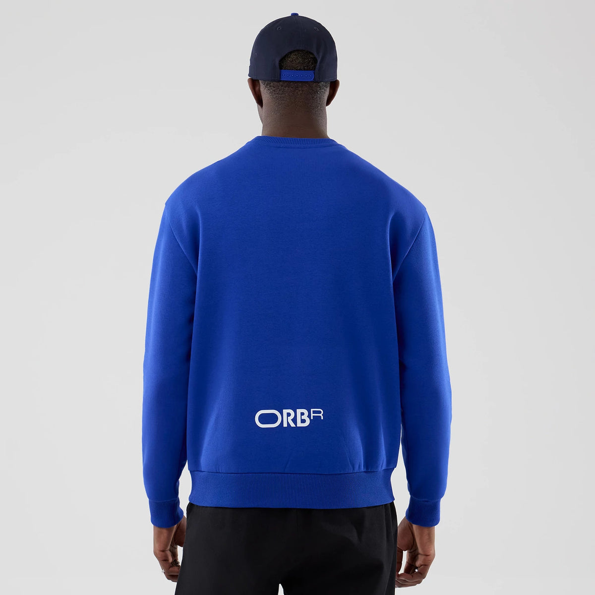 New Era Red Bull Racing Blue Oversized Crew Neck Sweater, back view on a model wearing a matching navy cap, featuring white ORBR lettering centred near the hem, ribbed cuffs and waistband, highlighting its blue fabric.
