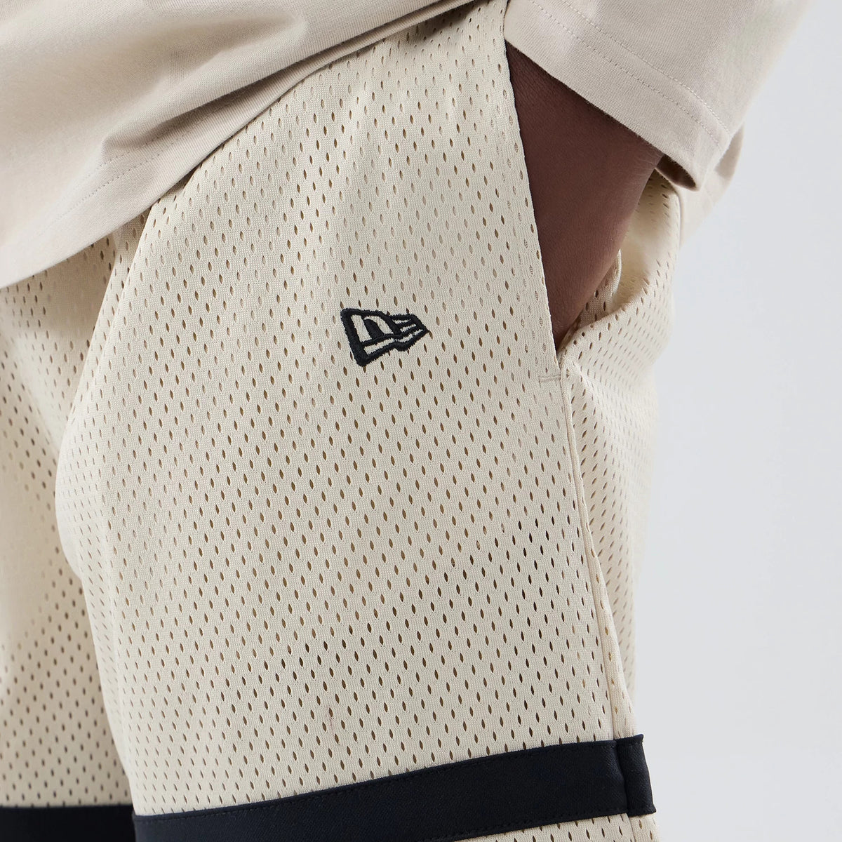 New Era New York Yankees MLB Light Beige Oversized Mesh Shorts, three-quarter view showing a black embroidered New Era logo on the front near the pocket, light beige mesh fabric, and a contrasting black band along the hem.