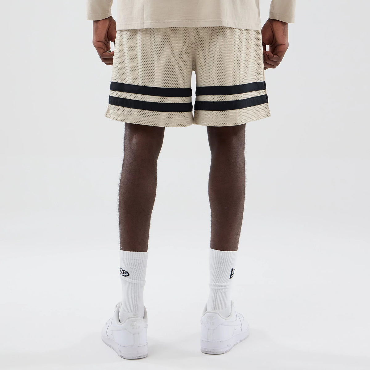 New Era New York Yankees MLB Light Beige Oversized Mesh Shorts, back view on a model, showcasing the light beige mesh fabric with double black horizontal stripes near the hem, contrasted against white sneakers.