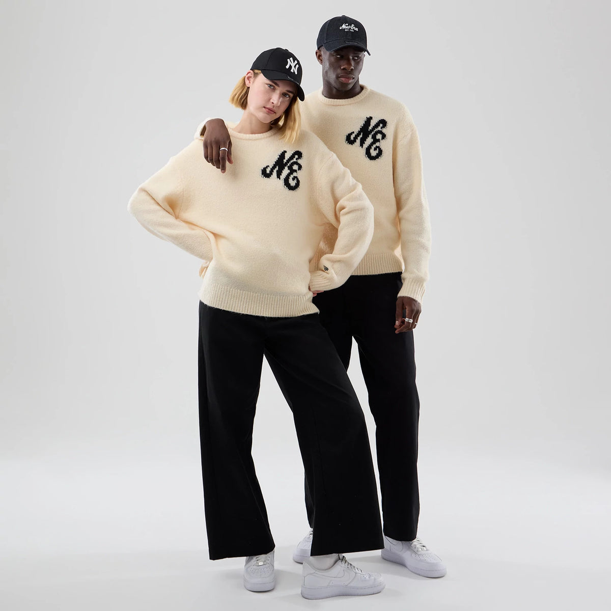 New Era Knit Cream Knitted Sweater, on-model view featuring a relaxed fit with black ‘N & C’ lettering on the chest, ribbed trims at the cuffs and hem, worn by two people in casual black trousers and white trainers.