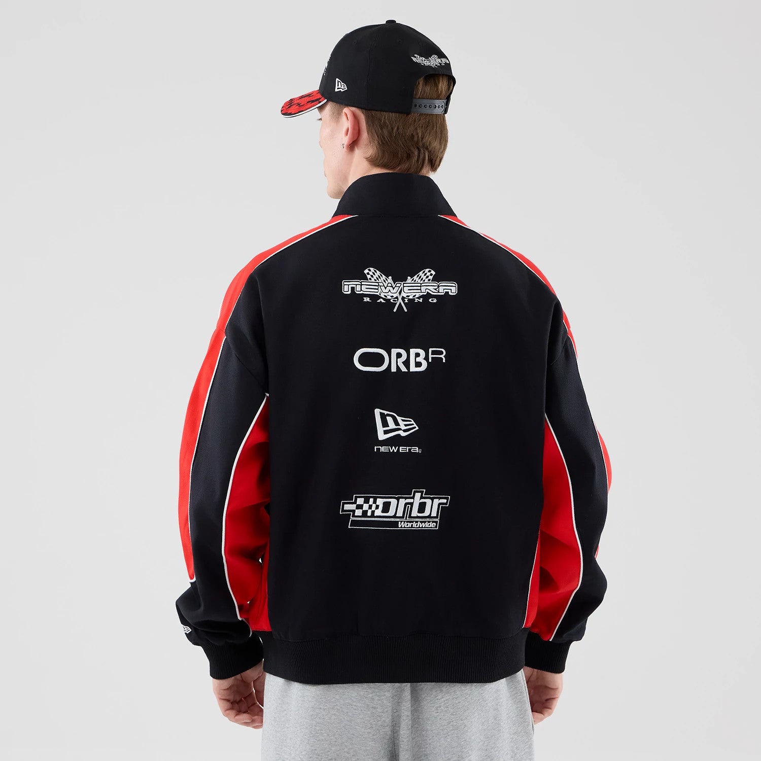 New Era Red Bull Racing Motorsport Black Jacket, back view on a model wearing a black cap, featuring multiple white embroidered logos including New Era, ORBR, and Red Bull Racing on the upper back, and red and white striped on the sleeves.