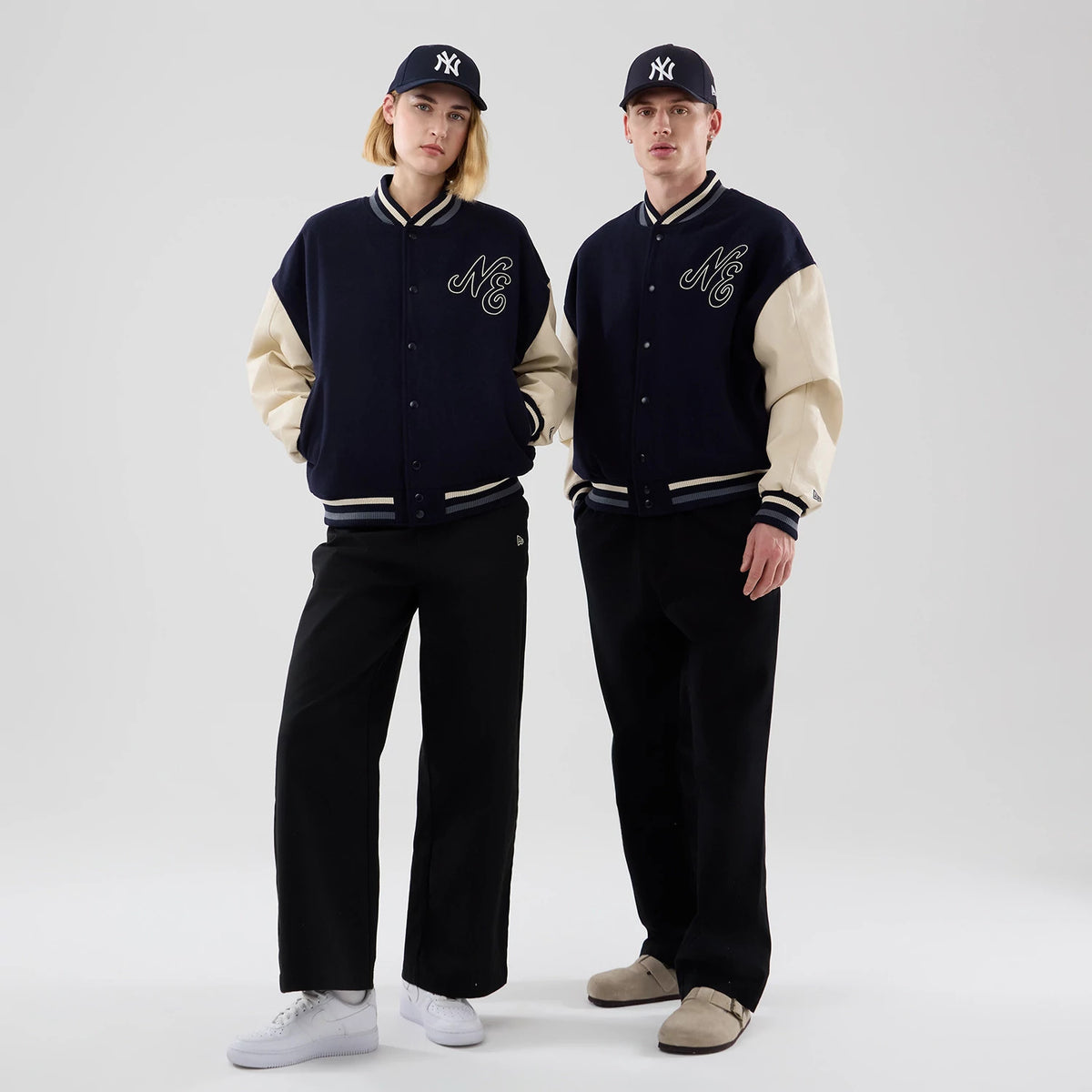 New Era Oversized Navy Varsity Jacket, front view on models wearing matching black trousers and navy caps, featuring cream-coloured sleeves, NE lettering embroidered on the chest, and striped trim details at the cuffs and waistband.
