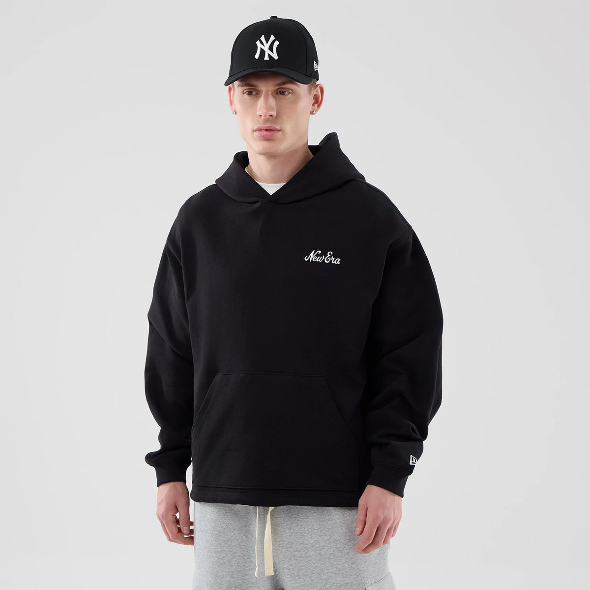 New Era Open Hem Black Pullover Hoodie, front view on-model showing the black hoodie with a white embroidered New Era logo on the left chest, large front pocket, and the New Era logo on the left sleeve above the cuff.