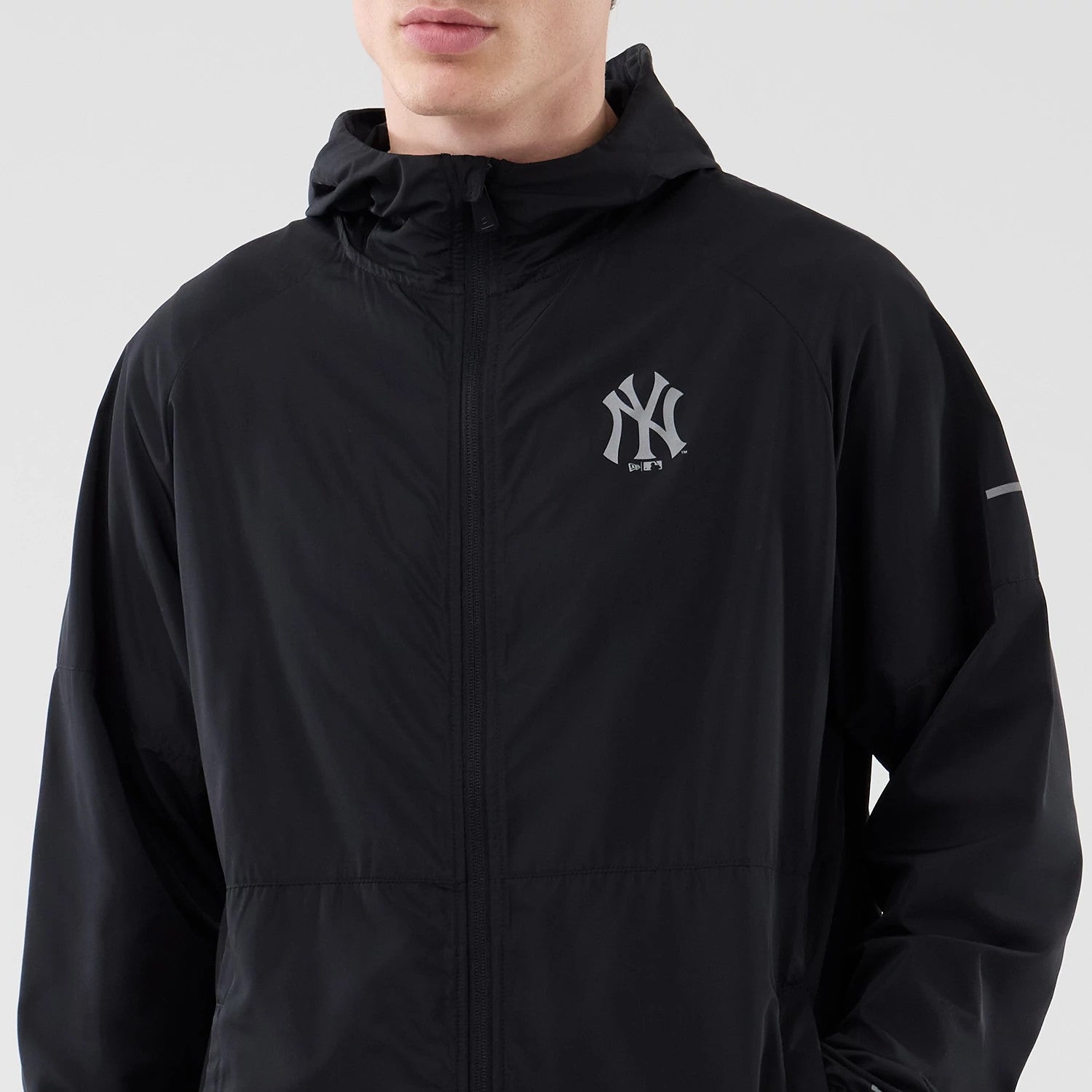 New Era New York Yankees MLB Core Plus Windbreaker Black Jacket, front view showing the black windbreaker with a printed New York Yankees logo on the left chest, a full zip closure up to a high collar, and a reflective stripe on the left arm.