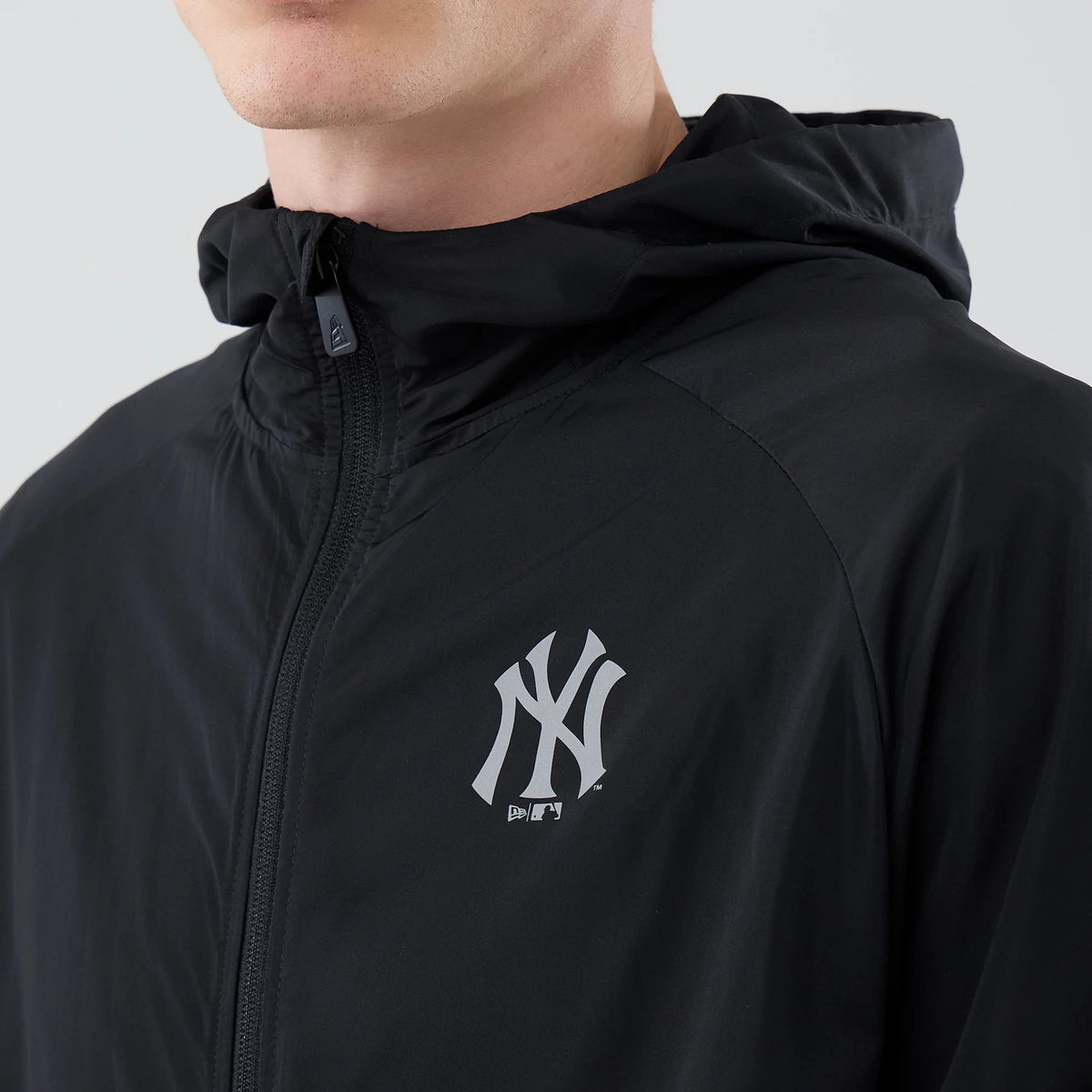 New Era New York Yankees MLB Core Plus Windbreaker Black Jacket, front view focusing on the silver printed Yankees logo on the left chest, featuring a full zip closure up to the hood, displayed on a person wearing the jacket with the hood down.