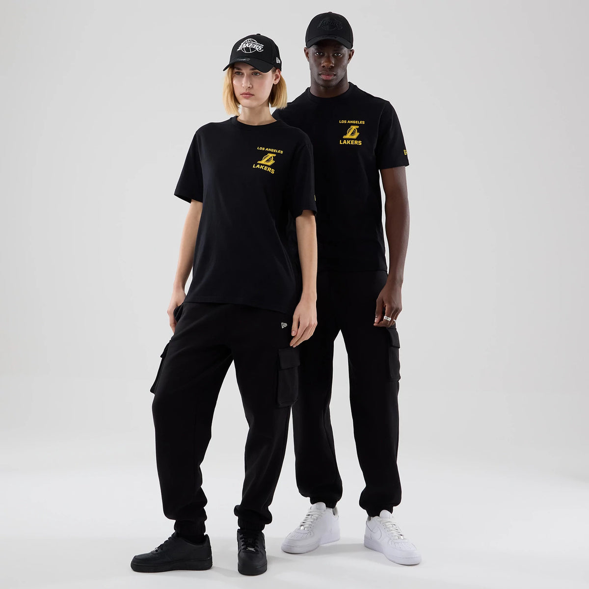 New Era LA Lakers NBA Court Graphic Black T-Shirt, front view on a model styled with cargo trousers and a cap, displaying a yellow printed Lakers logo and Los Angeles Lakers text on the left chest and another logo on the left sleeve.