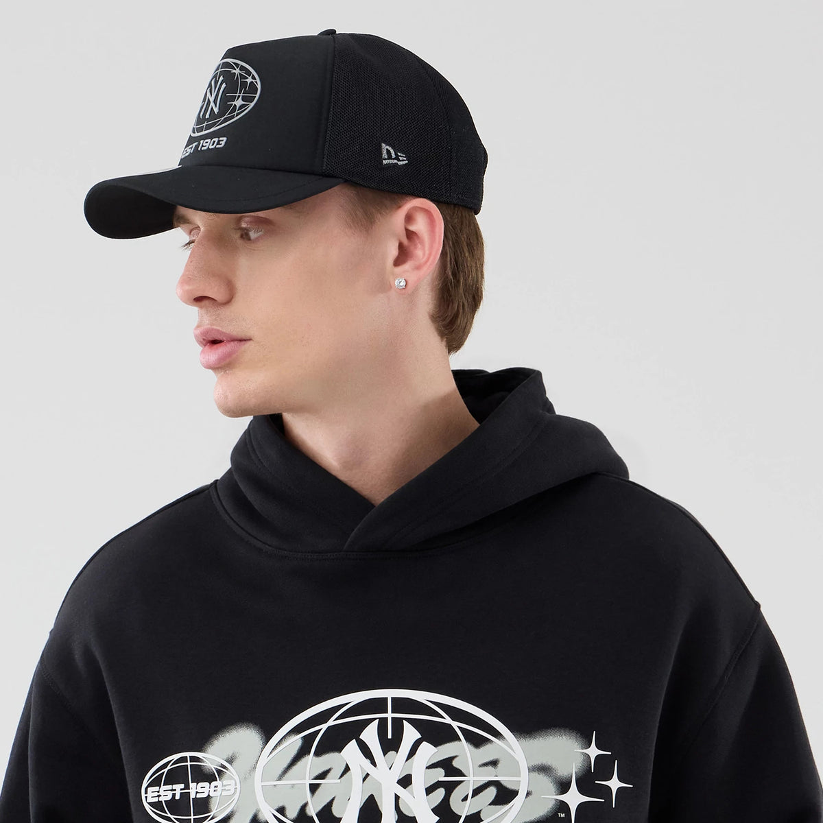 New Era New York Yankees MLB Logo Graphic Black Oversized Pullover Hoodie, on-model view showing the large white graphic with the Yankees logo on the chest, complemented by a black cap featuring an embroidered front circle logo and side logo.