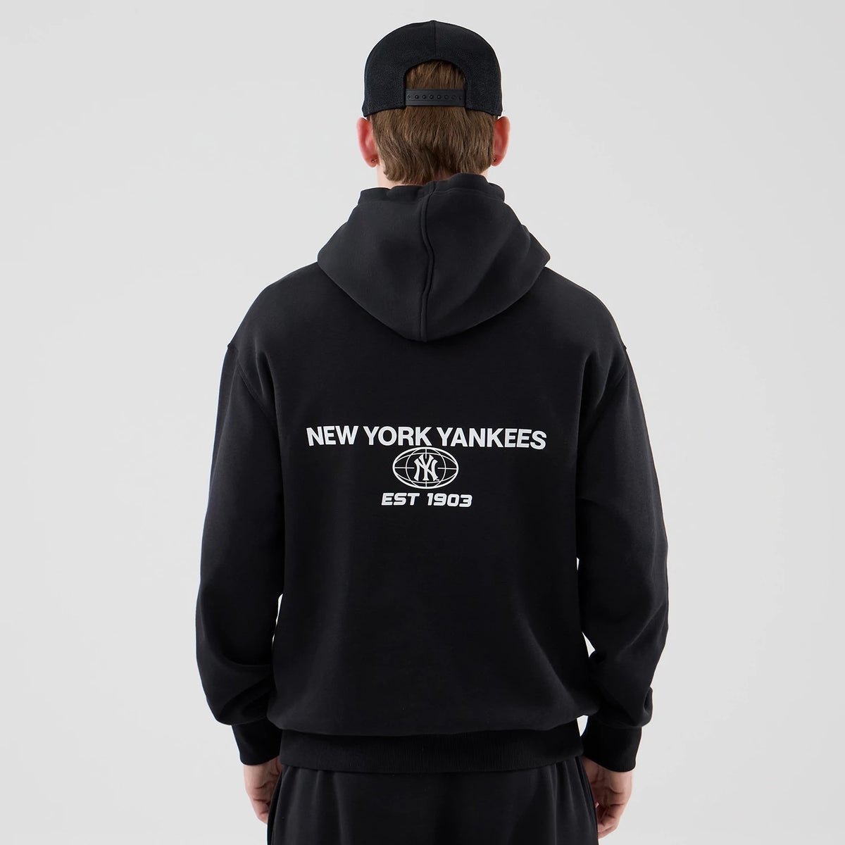 New Era New York Yankees MLB Logo Graphic Black Oversized Pullover Hoodie, back view on a model wearing a black cap, featuring the white embroidered New York Yankees text across the upper back above the NY logo and EST 1903 beneath it.