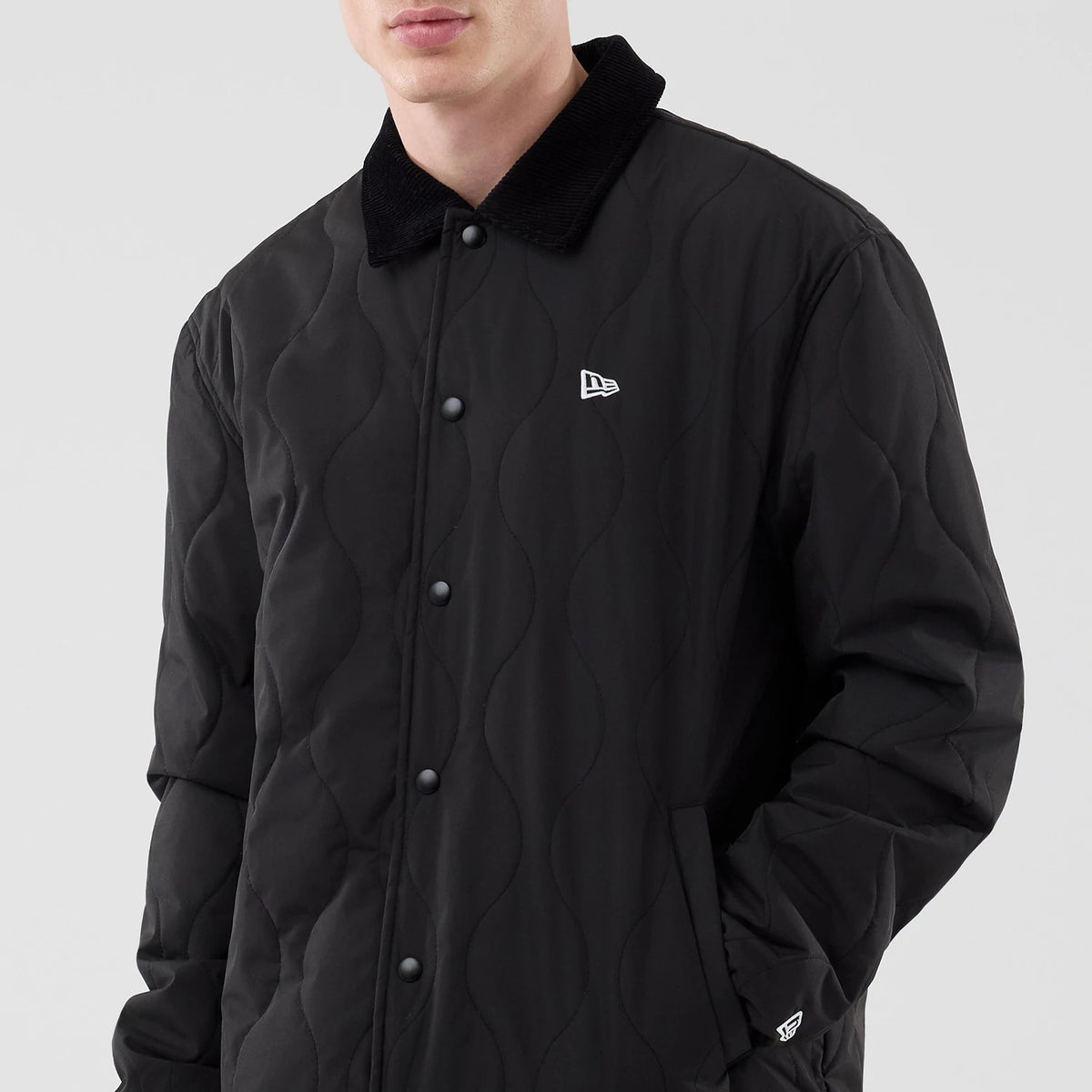 New Era Quilted Coaches Black Jacket, front view on a model highlighting the wavy quilted pattern across the black fabric, an embroidered New Era logo on the left chest, a button-down closure, and contrasting collar.