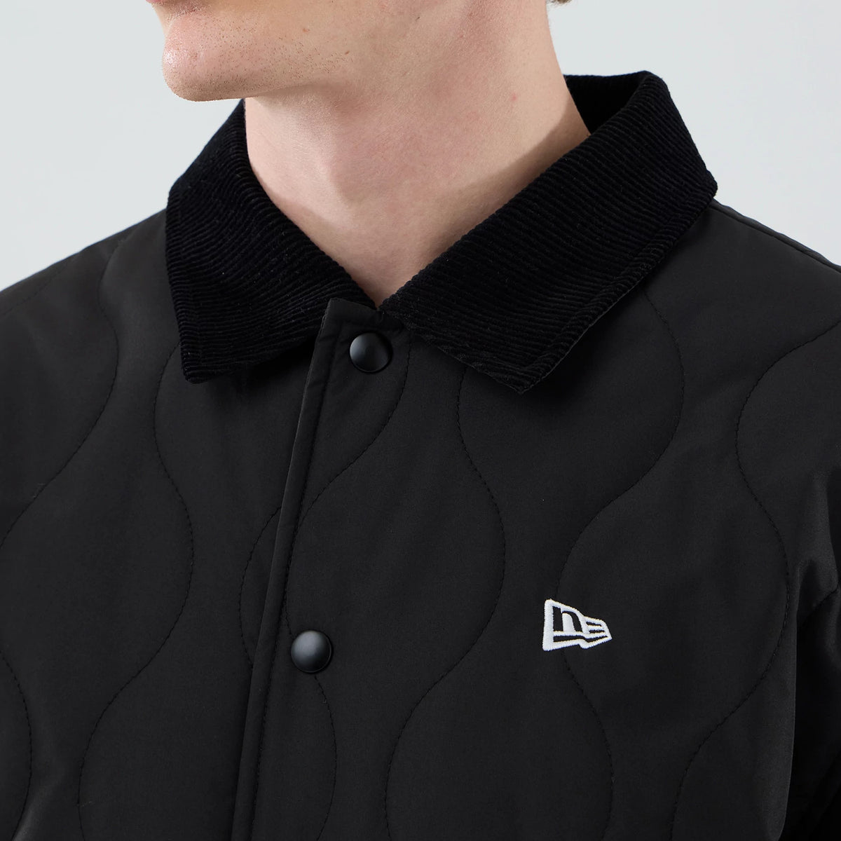 New Era Quilted Coaches Black Jacket, front view on a model highlighting the textured black quilted pattern, a contrasting corduroy collar, and the embroidered New Era logo in white on the left chest near the matte black button closure.