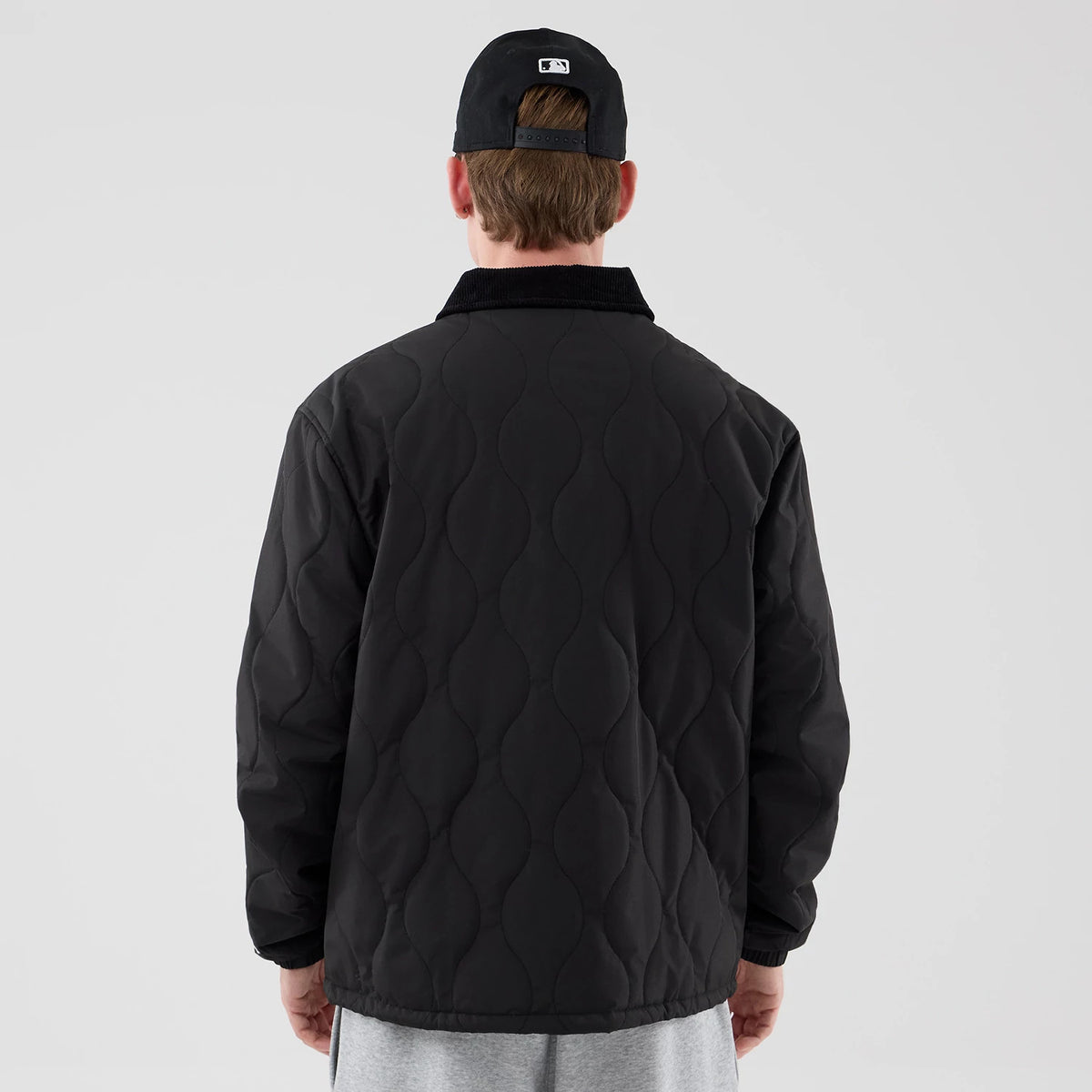 New Era Quilted Coaches Black Jacket, back view on a model showing the distinctive wavy quilted pattern across the back, a solid black collar, and pairing with a black cap worn backwards featuring an embroidered logo above the closure.