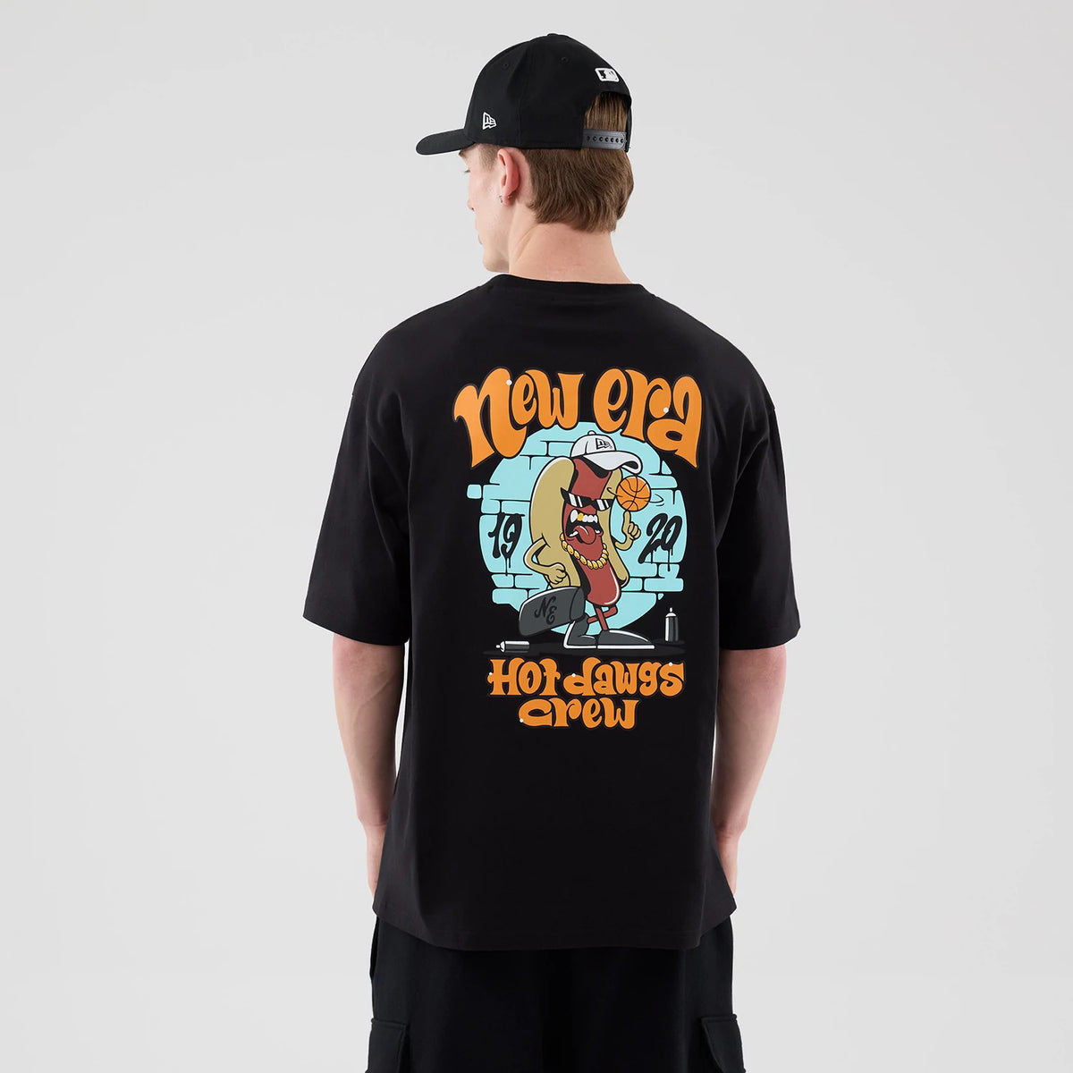 New Era Hot Dog Graphic Black Oversized T-Shirt, back view on a model showing a colourful graphic of a hot dog character wearing a cap, holding a basketball, with New Era lettering above and Hot Dawgs Crew text below.