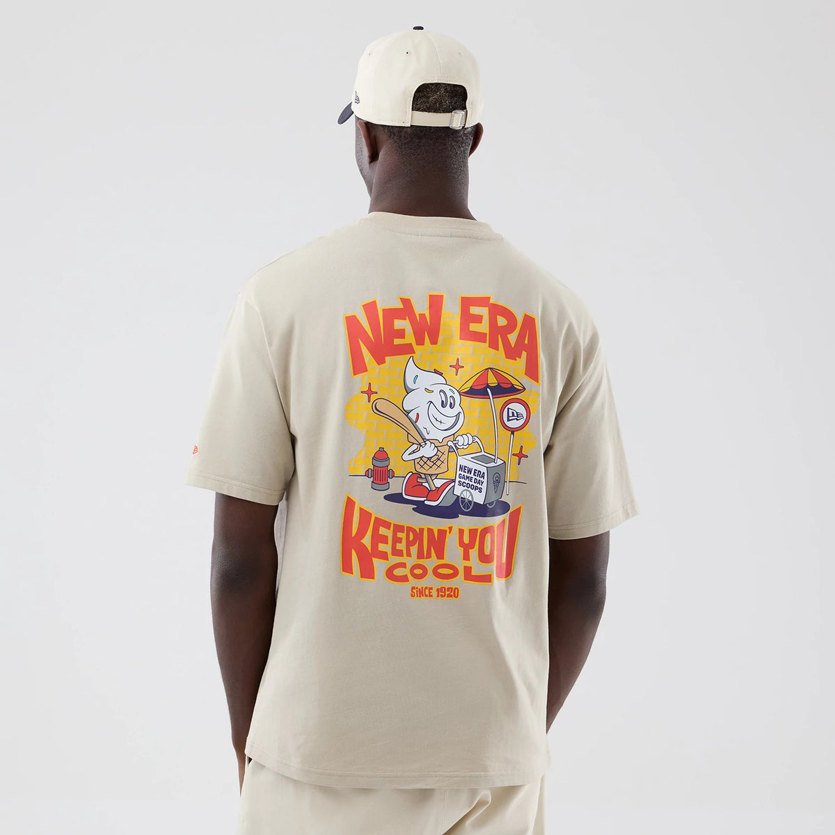 New Era Food Graphic Light Beige Oversized T-Shirt, back view on a model featuring a colourful cartoon design with an ice cream character, New Era logo on an umbrella, and Keepin' You Cool lettering below.