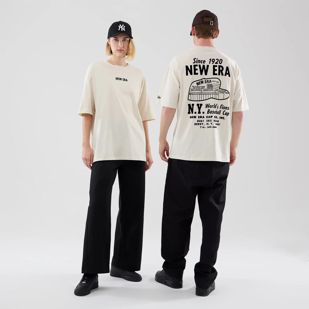 New Era Cap Graphic Cream Oversized T-Shirt, on-model view showing front and back designs with small printed New Era lettering on the chest, large vintage-inspired graphic and text across the upper back, styled with black trousers and caps.
