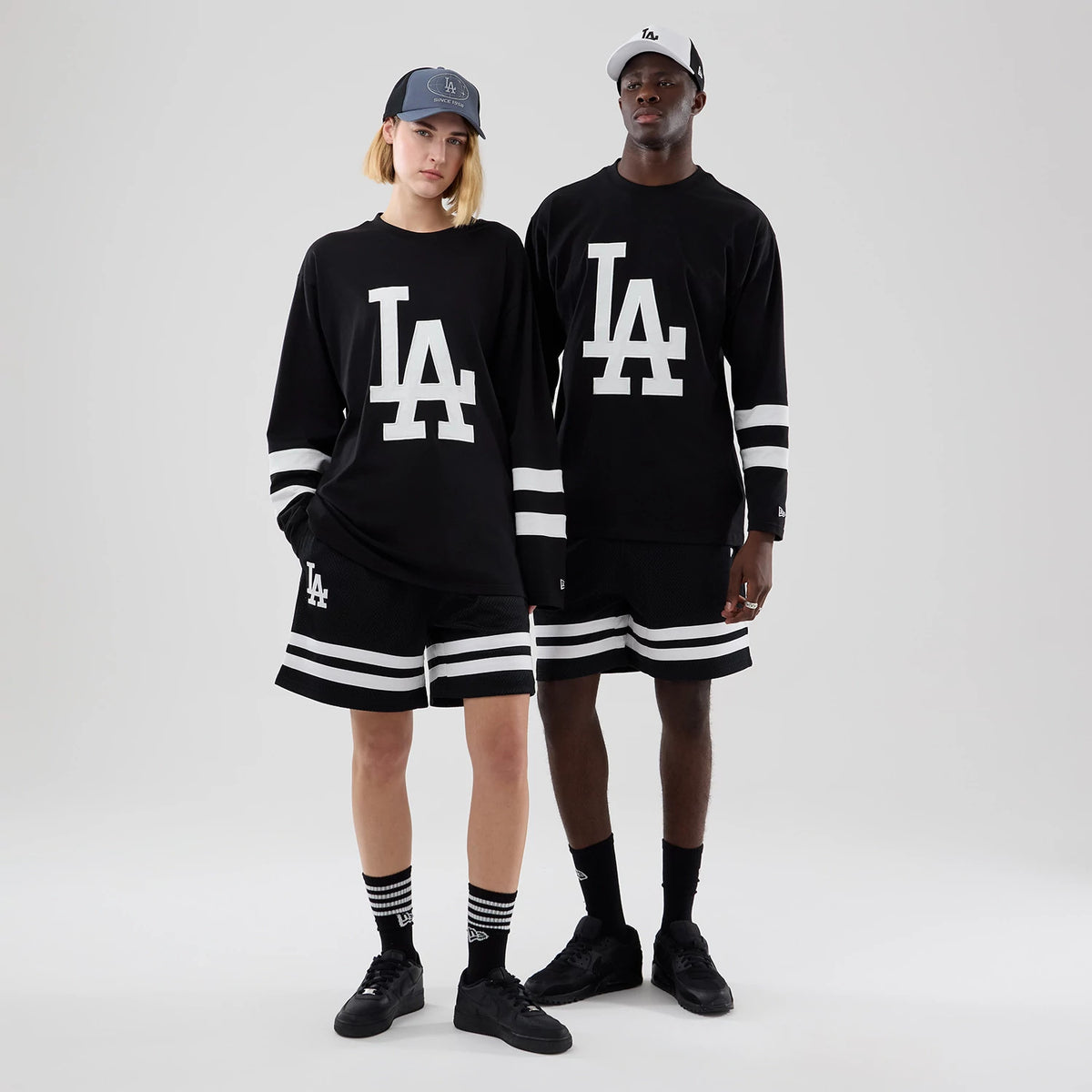 New Era LA Dodgers MLB Black Oversized Long Sleeve T-Shirt, on-model view featuring a large white LA logo centred on the chest, horizontal white stripes on the sleeves, with a model wearing matching black shorts with white stripes and a grey cap.