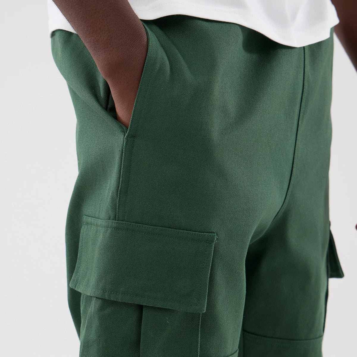 New Era Dark Green Cargo Trousers, side view showing a close-up of the cargo pocket with a flap on the left thigh, a hand casually placed in the side pocket, and the texture of the dark green fabric.