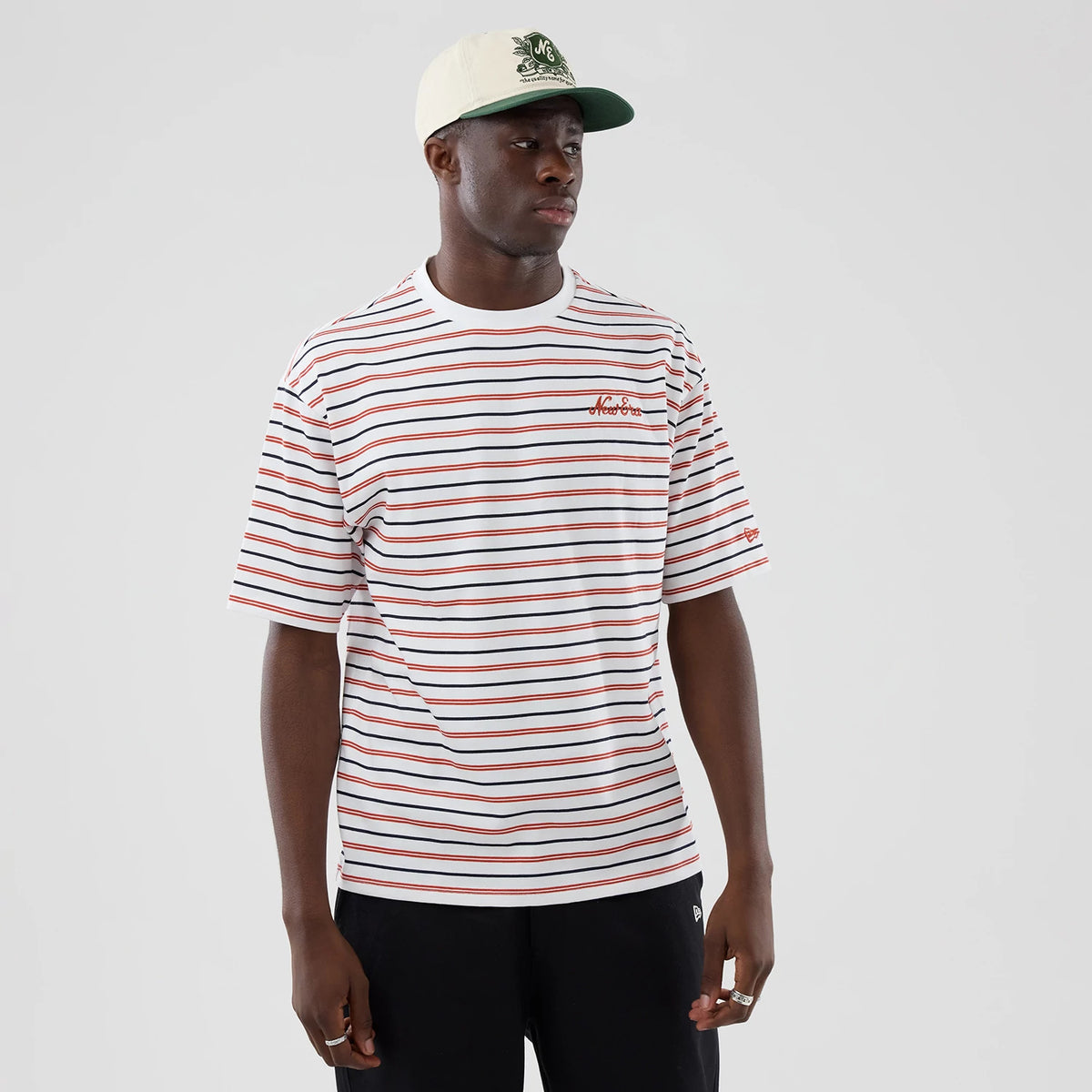 New Era Stripe White Oversized T-Shirt, front view on a model wearing a light cap, featuring horizontal red and black stripes across the white body, an embroidered script logo on the left chest, and a small logo on the left sleeve edge.