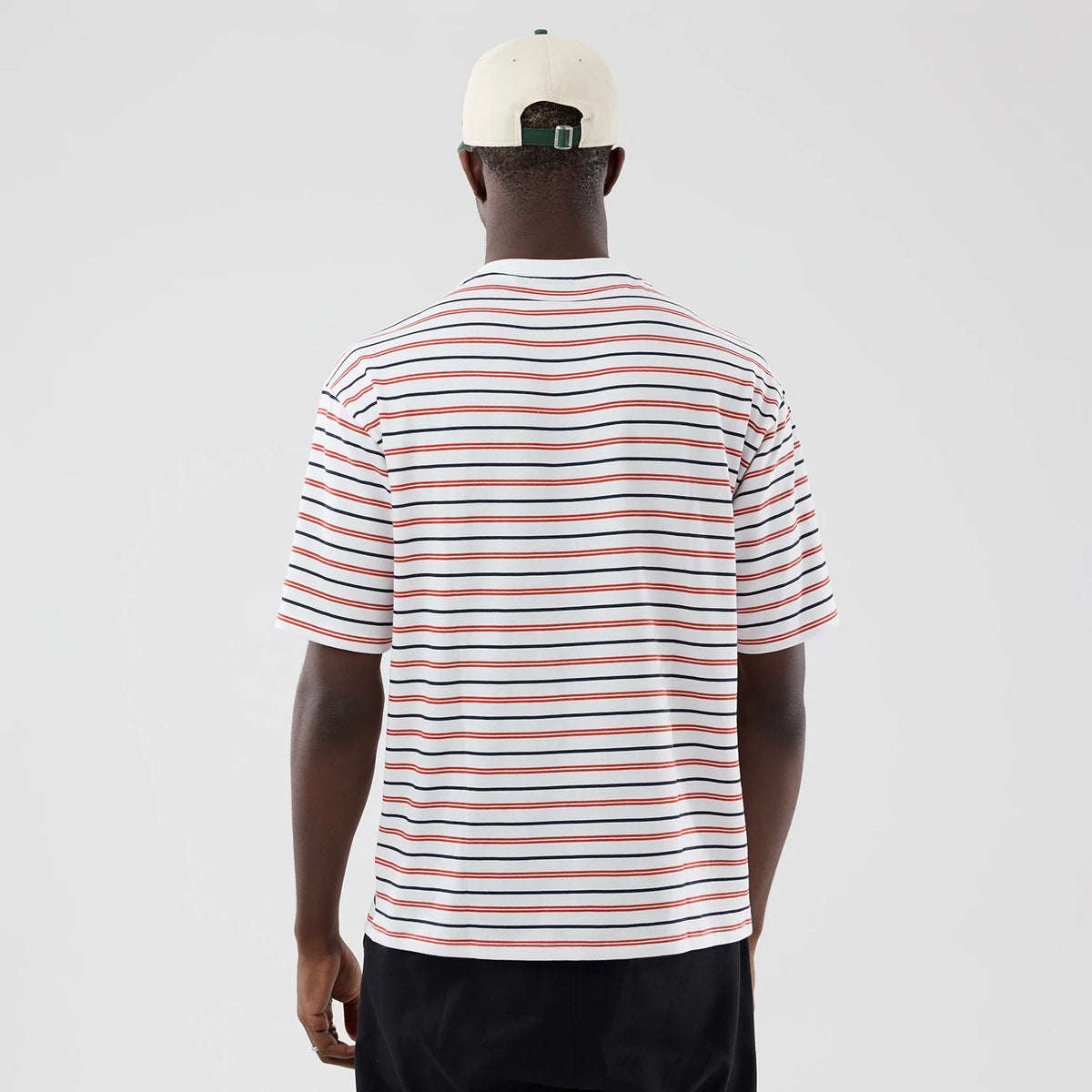 New Era Stripe White Oversized T-Shirt, back view on a model showcasing the horizontal stripes in white, black, and red across the shirt, short sleeves and a relaxed fit, worn with a neutral cap and dark trousers.
