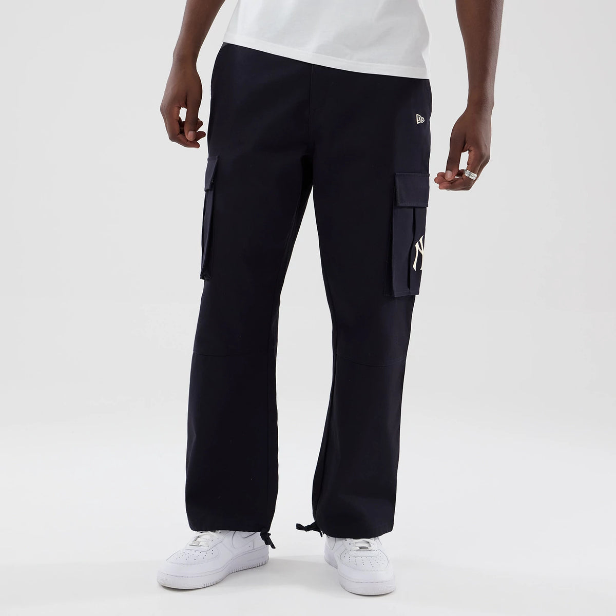 New Era New York Yankees MLB Navy Cargo Trousers, front view on a model showing the embroidered New Era flag logo on the left thigh pocket, the white NY logo on the right pocket, and adjustable drawstring cuffs above white trainers.