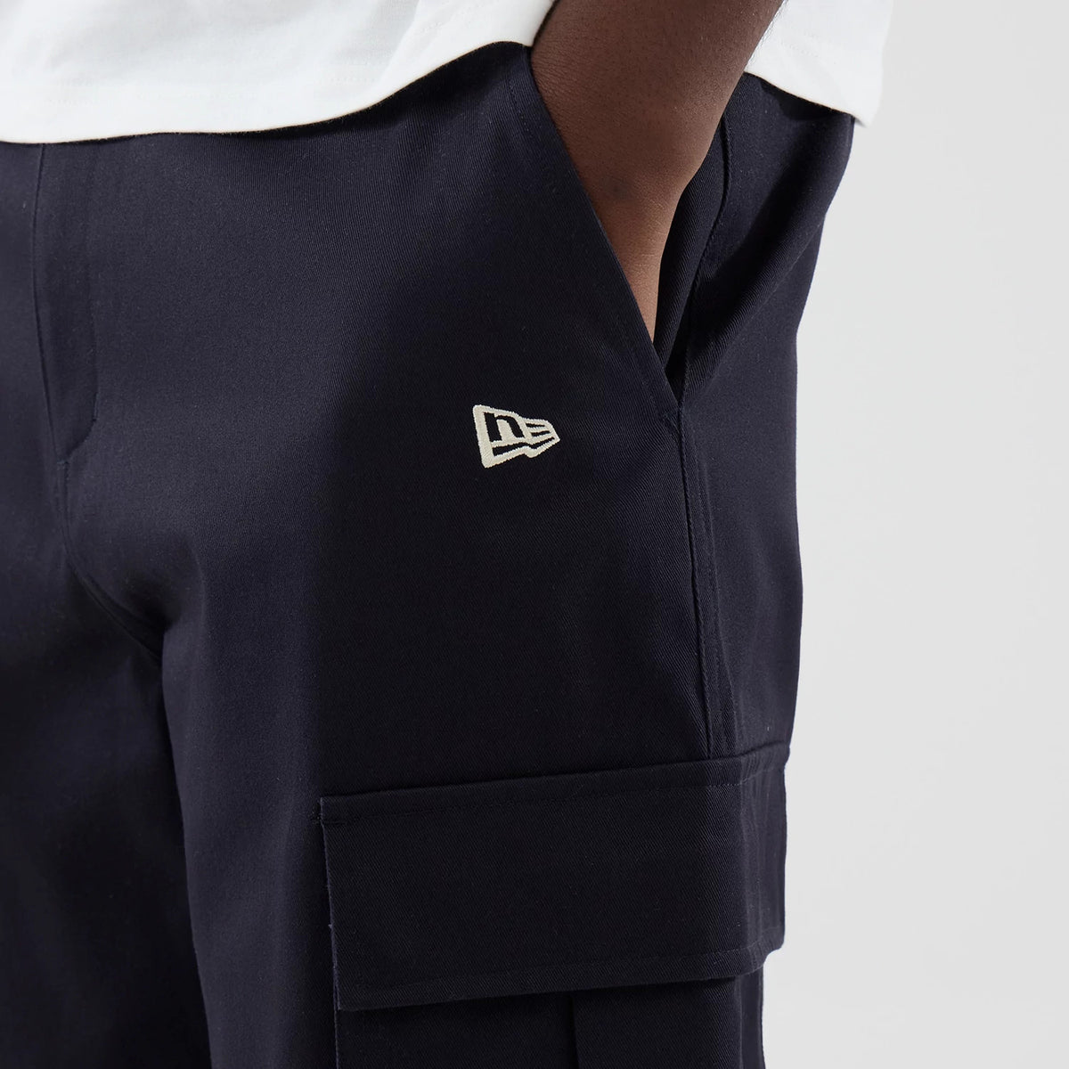 New Era New York Yankees MLB Navy Cargo Trousers, close-up view showing the New Era flag logo embroidered in white on the left thigh, with deep side pockets and an additional pocket on the side of the leg, styled on a model wearing a white top.