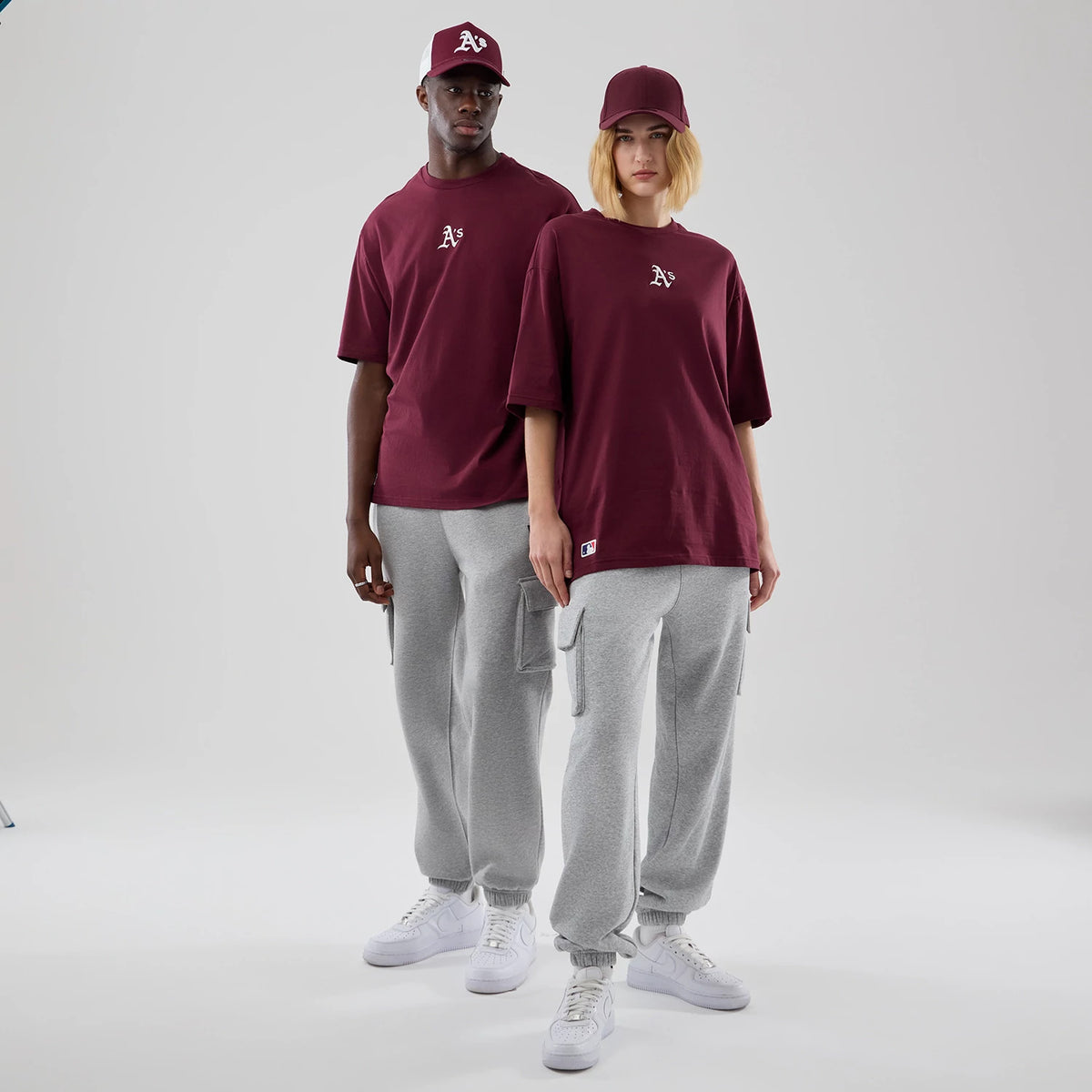 New Era Athletics MLB Midi League Essential Dark Red Oversized T-Shirt, front view on a model styled with grey joggers and white trainers, showcasing the white embroidered A's logo on the chest and the relaxed fit of the dark red tee.
