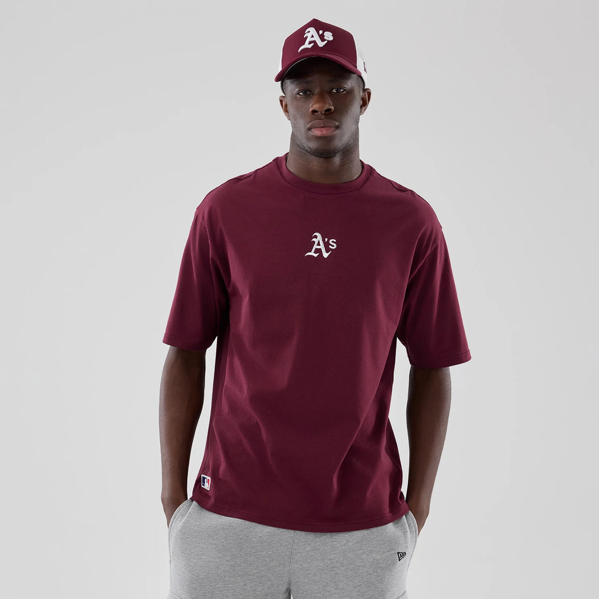 New Era Athletics MLB Midi League Essential Dark Red Oversized T-Shirt, front view on-model showing the dark red shirt with a white embroidered A's logo centred on the chest, paired with a matching cap and grey shorts featuring a New Era flag.