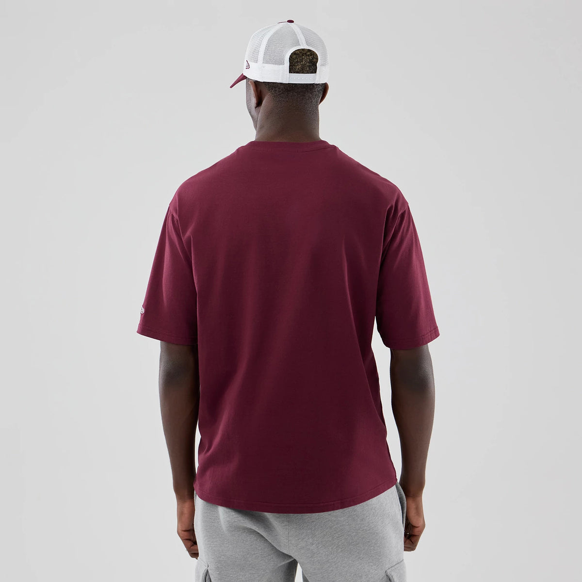 New Era Athletics MLB Midi League Essential Dark Red Oversized T-Shirt, back view on a model wearing grey shorts and a white mesh cap, showing a plain dark red back with a small New Era embroidered logo on the left sleeve.