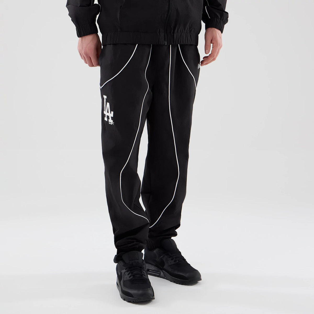 New Era LA Dodgers MLB Piping Track Black Track Pants, front view on a model with white piping curving along the thighs and down the legs, a white LA logo embroidered on the right leg, and elasticated cuffs above black trainers.