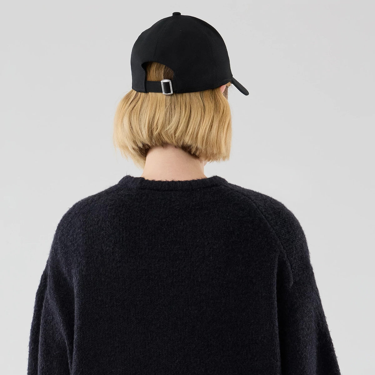 New Era Knit Heritage Navy Knitted Sweater, rear view on a model with a black baseball cap, showing the textured knit pattern and relaxed fit of the navy sweater, complementing the adjustable strap and buckle of the cap.