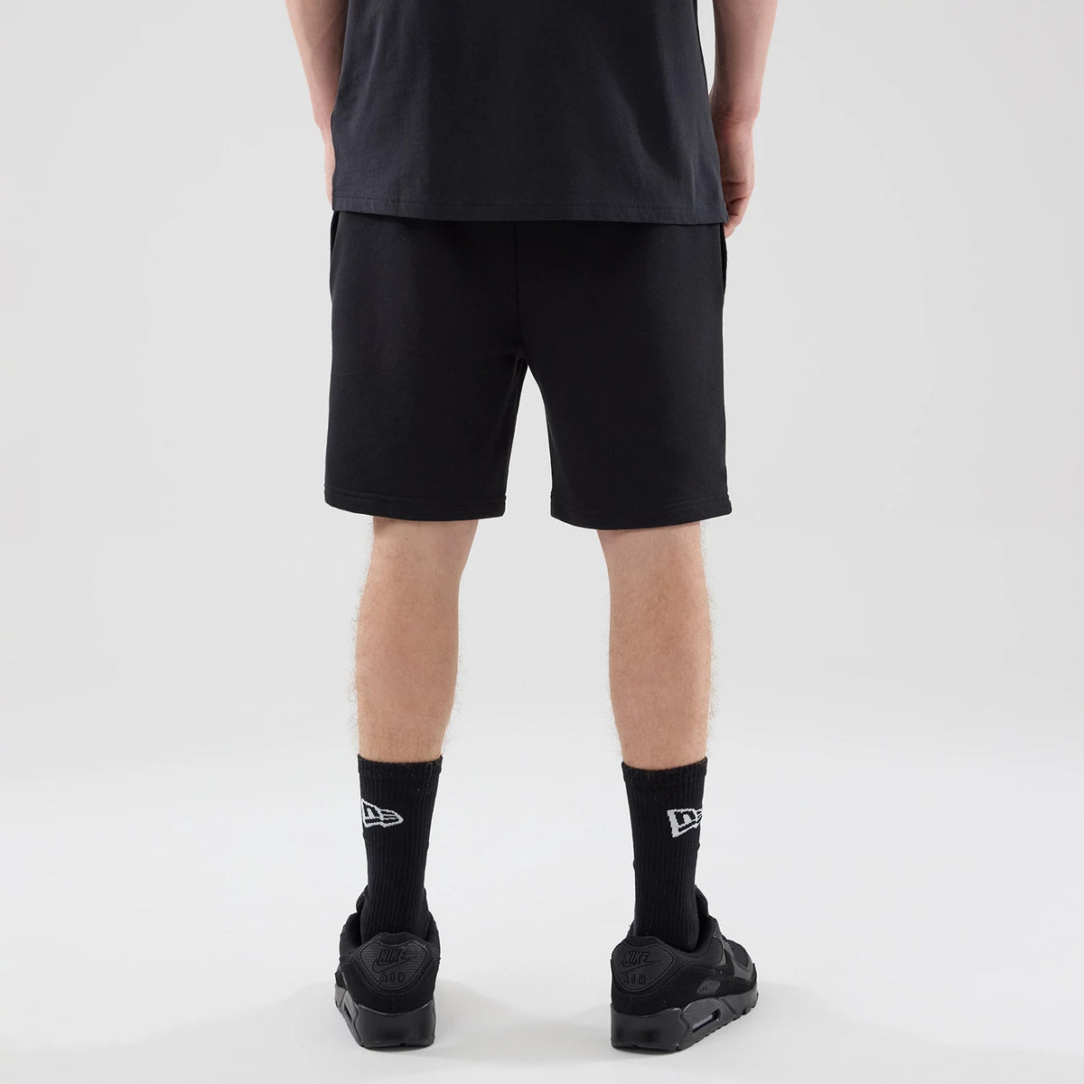 New Era Chicago Bulls NBA League Essential Black Oversized Shorts, back view on-model showing the plain black design extending to just above the knee, with elastic waistband for relaxed fit, paired with New Era branded socks and black trainers.