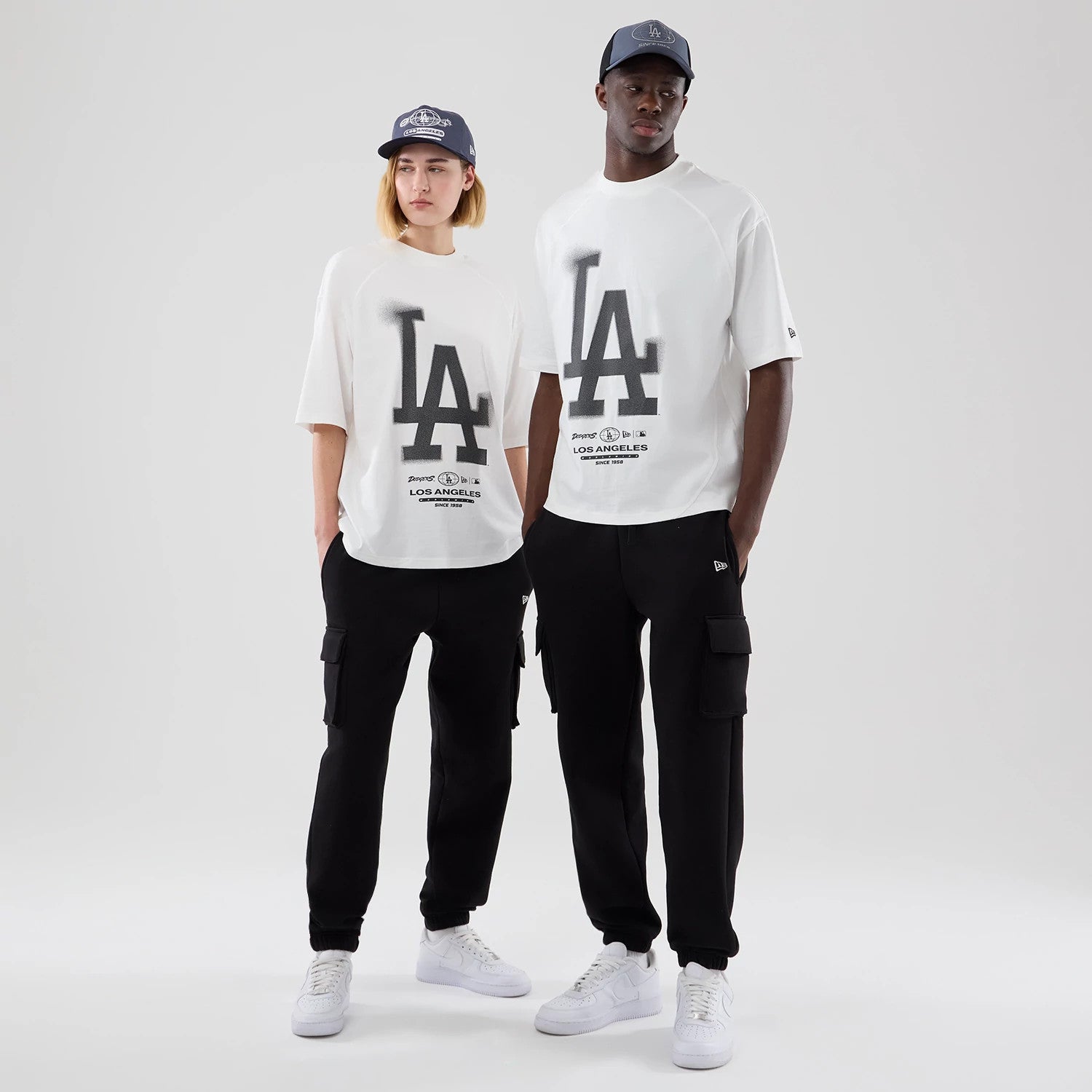 New Era LA Dodgers MLB Graphic White Boxy T-Shirt, front view on-model showing the large black LA logo spanning the chest, smaller team graphics below, and styled with black trousers and white trainers, both models also wearing matching caps.