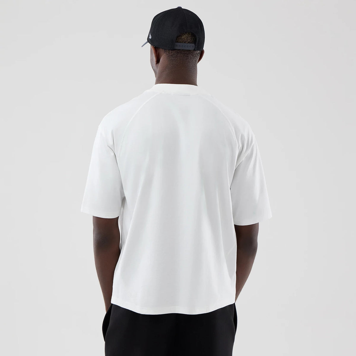 New Era LA Dodgers MLB Graphic White Boxy T-Shirt, back view on a model with a black cap, highlighting the wide fit and plain white back with raglan sleeves, creating a clean and minimalistic appearance against dark trousers.
