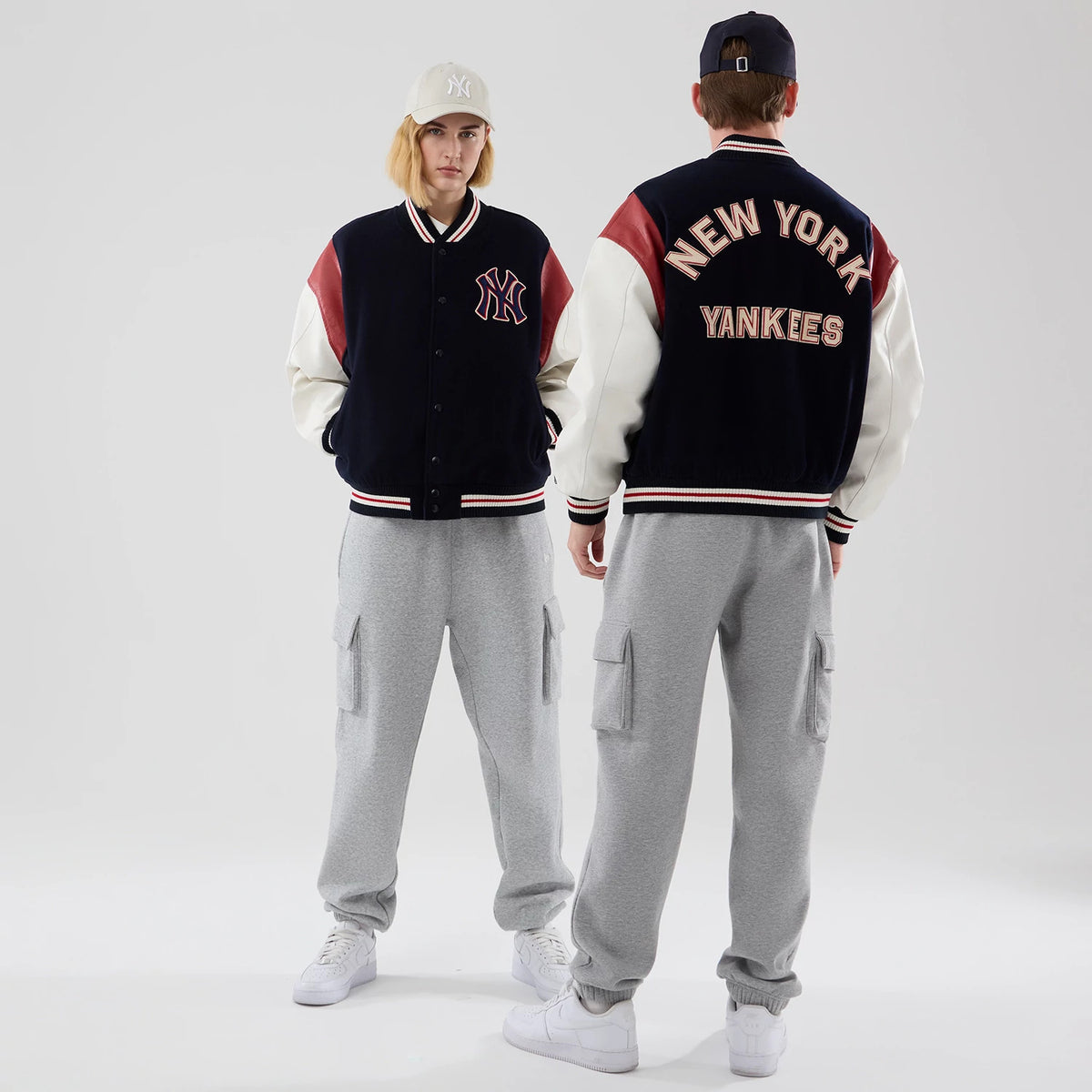 New Era New York Yankees MLB Varsity Navy Jacket, front and back view on models with caps, showing the NY logo embroidered on the left chest, red and white sleeve stripes, and New York Yankees lettering across the back shoulders.