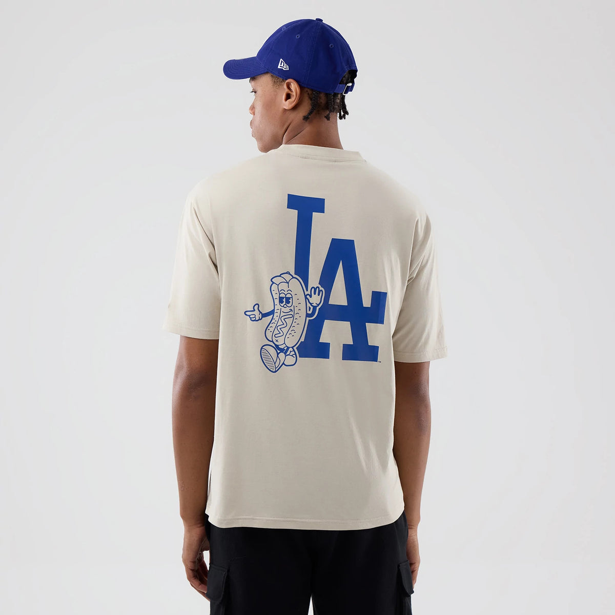 New Era LA Dodgers MLB Food Graphic Cream Oversized T-Shirt, back view on a model with a blue cap, showing a large blue printed LA Dodgers logo on the centre back, accompanied by a playful hot dog character graphic pointing to the side.